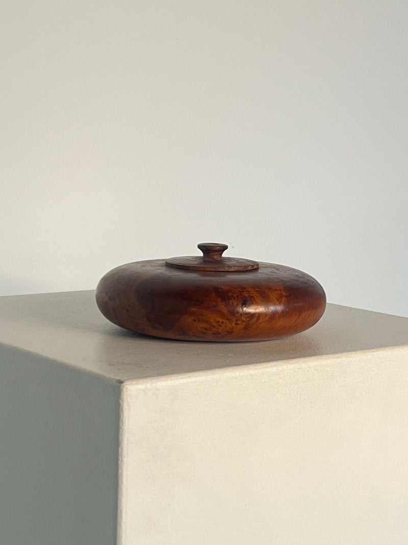 The **Walnut Burl Shallow Bowl With Lid** by **Porter Studio** sits elegantly on a white pedestal against a plain background. Its round, polished form features a small knob, while the bowl's rich brown hue and visible grain patterns enhance its refined appearance.