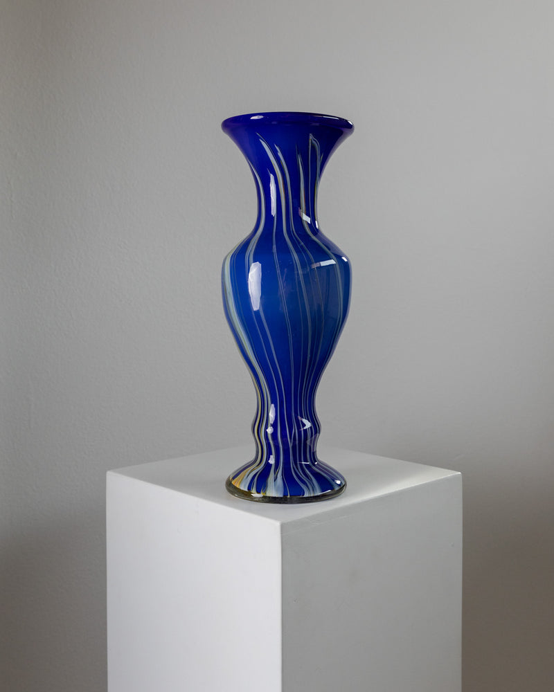 A Murano Glass Vase Blue by Bottega Jacobs, from 1970s Italy, is showcased on a white pedestal against a light gray background, highlighting exquisite Italian glass art with elegant white swirl patterns.