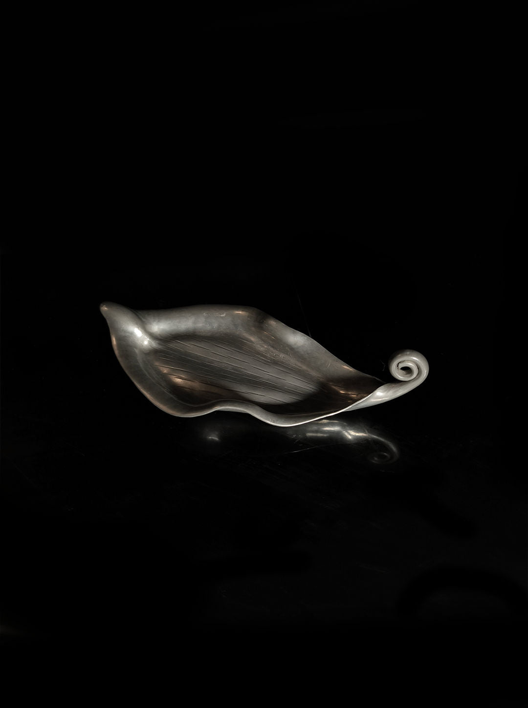 A silvery, metallic bowl from Les Objoies, called the Handmade Leaf Vide Poche, rests against a black background. This pewter decorative tray features a smooth, reflective surface with elegant, flowing curves that evoke an Art Nouveau vide-poche.