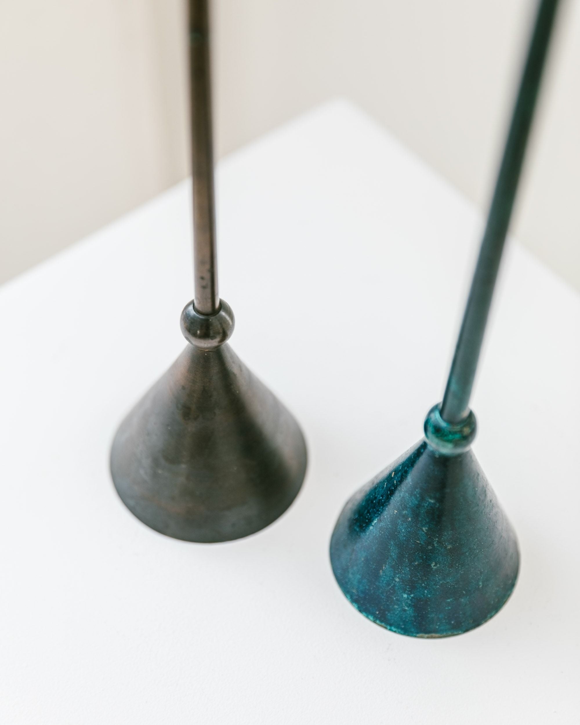 A set of two Bottega Jacobs candleholders stands on a white surface, showcasing a postmodern 1980s aesthetic. One features a dark brown cone with an attached metallic rod, while the other displays a teal tone; both have a slightly textured appearance that echoes their vintage charm.