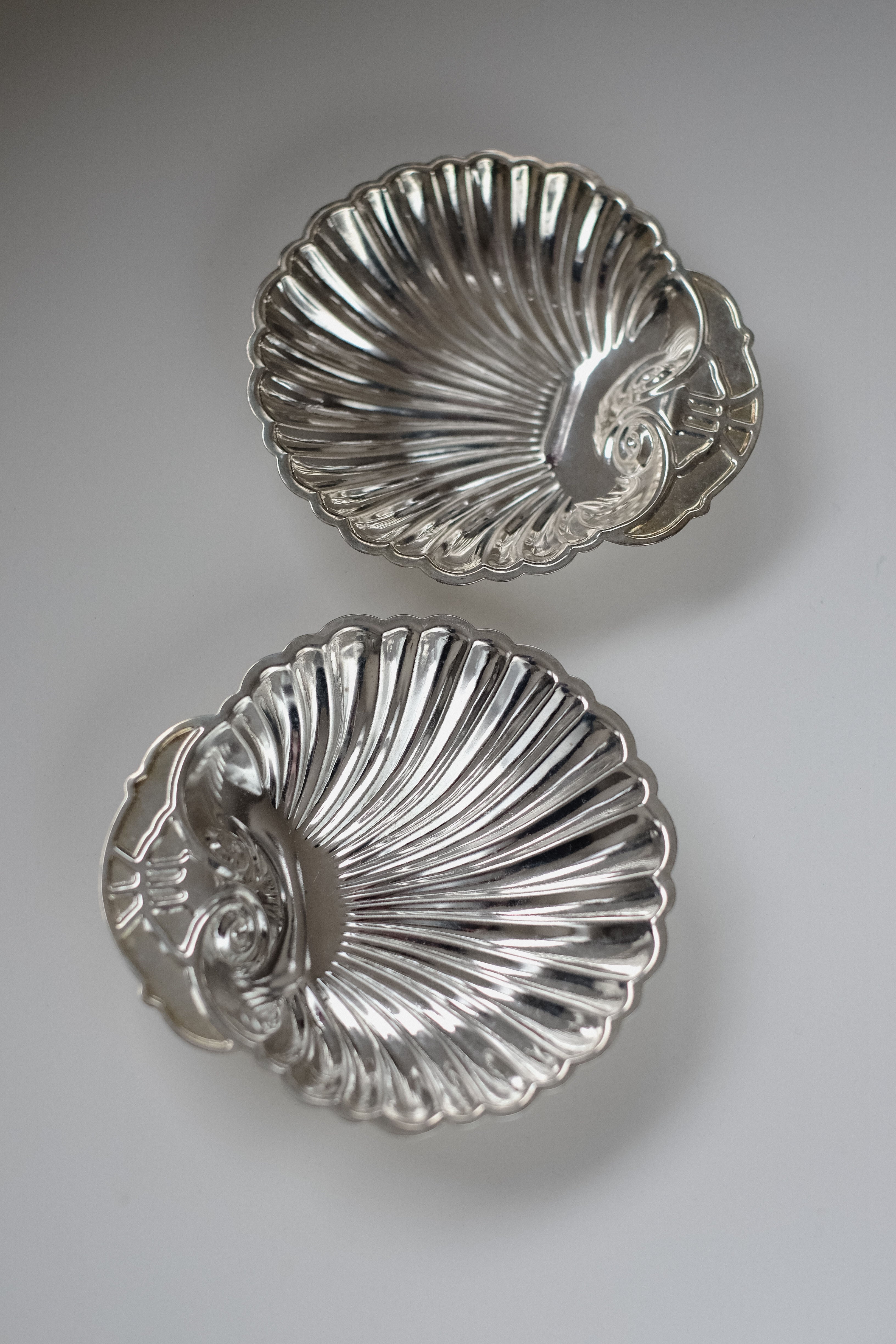 Displayed on a plain white surface, the Set of 2 Silver-plated Shell Trays by septembre studios features vintage shell shapes with ornate handles and a polished finish. Intricately detailed outer edges enhance their elegance, making them perfect for elevating any dining experience.