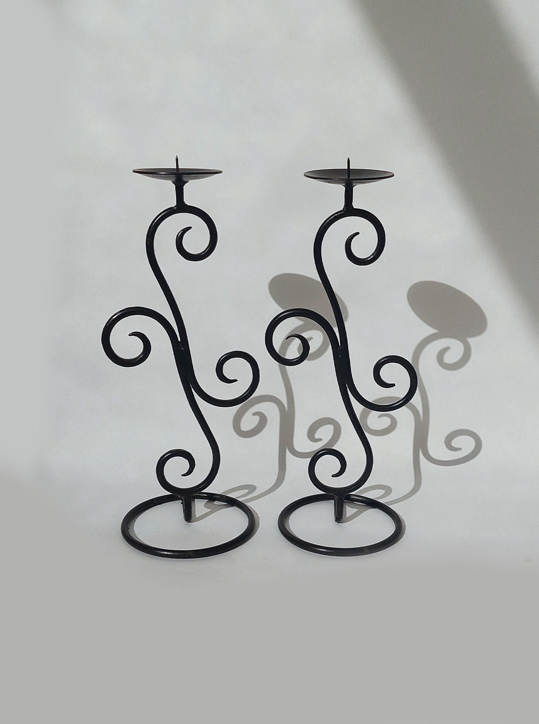 The Pair of Candle Holders by Les Objoies feature two black, wrought-iron pieces with intricate spiral designs standing on round bases, casting delicate shadows on a plain light background. Perfect for adding an elegant touch to your home décor.