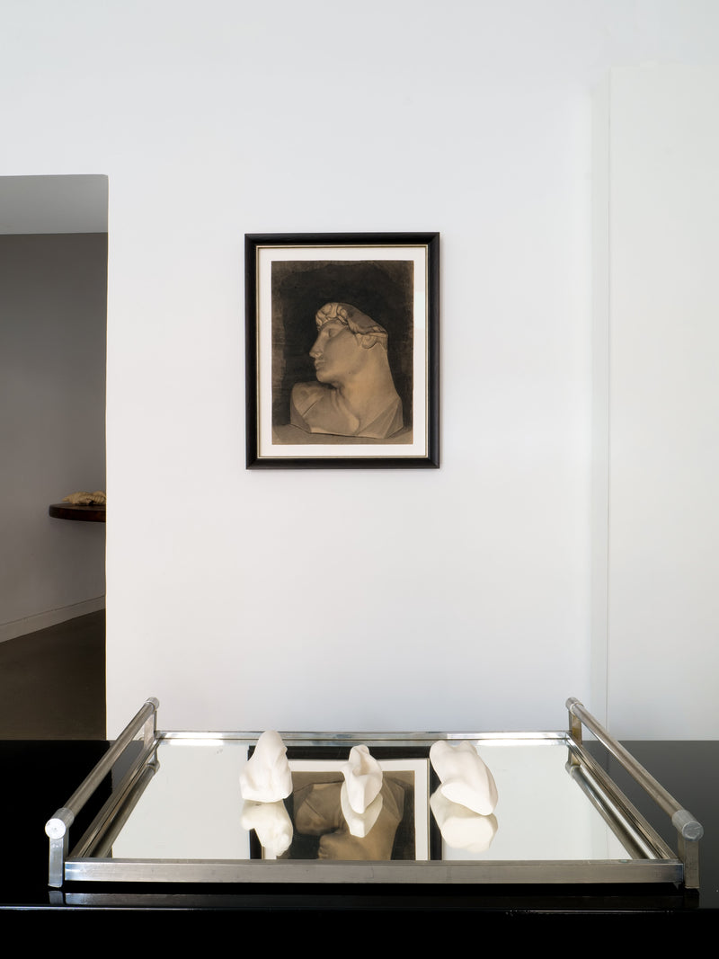 A modern art gallery wall showcases a framed piece, "Charcoal Sketch 1894" by Collection apart, depicting a person with their head slightly turned. Below this artwork sits a sleek black table with a reflective surface that displays three abstract white sculptures reminiscent of Michelangelo's masterpieces.