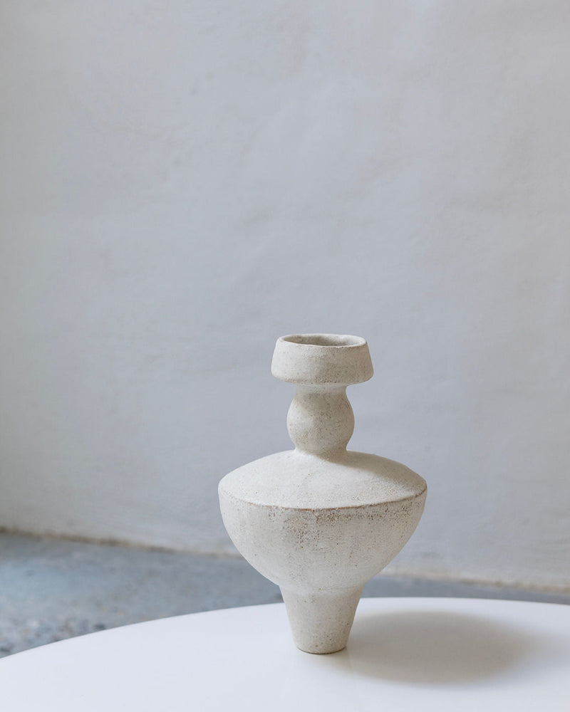 The Lydion Blanco Hueso ceramic vase by Canoa Lab features sculptural geometric forms and an off-white finish, displayed on a white surface with a light gray textured wall, evoking Mediterranean design inspired by ancient artifacts.