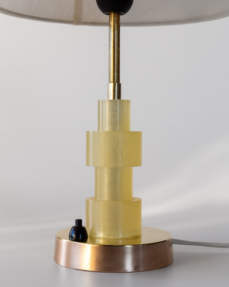 The Spigel Swedish Mid-Century Table Lamp features an Art Deco-style with a tiered yellow glass base, gold metal stem, and circular base with an on/off switch. Its textile shade peeks from the top, all set against a neutral backdrop.
