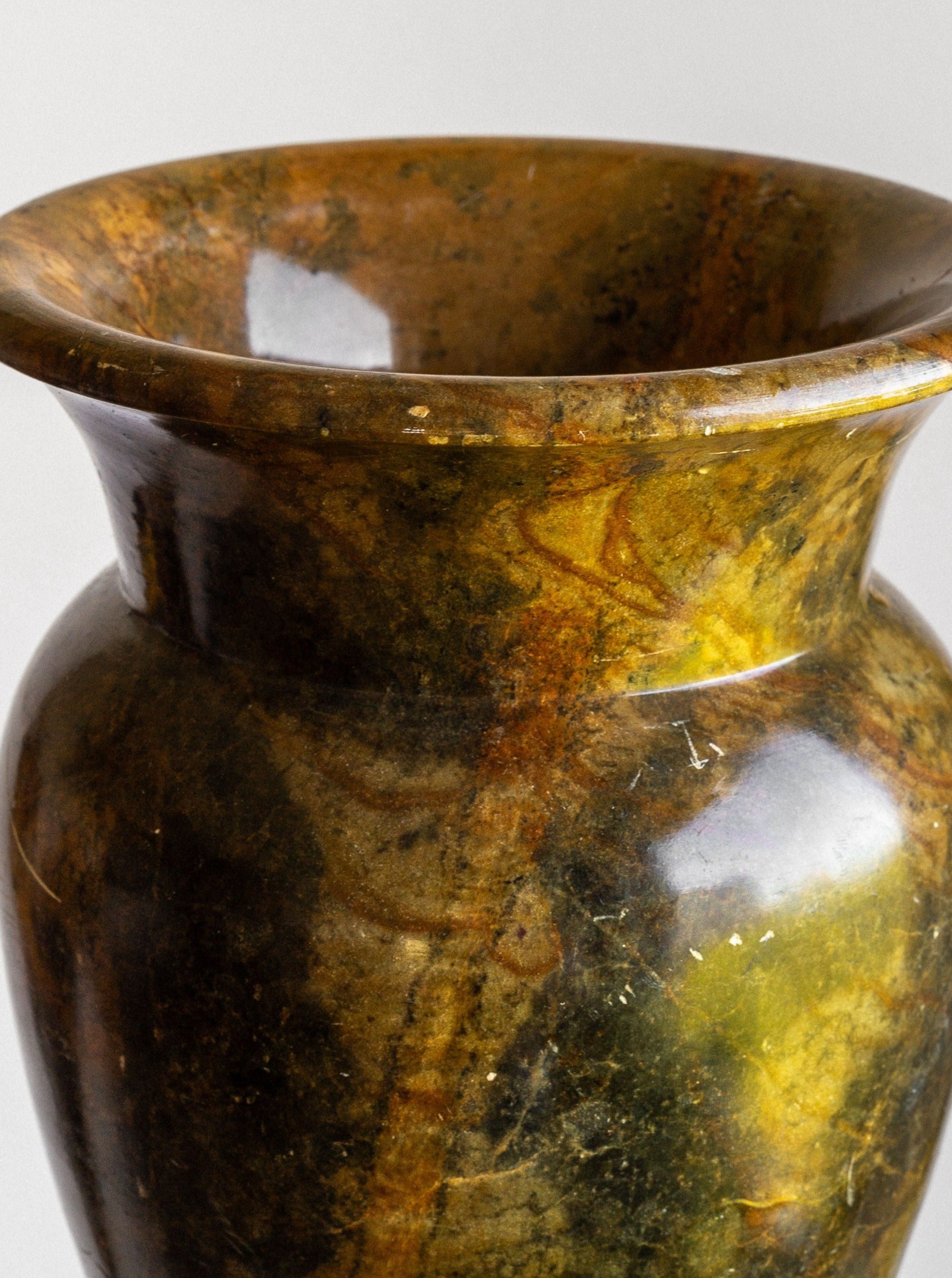 A close-up of the Spigel Large Neoclassical Stone Vase, featuring a green and yellow marbled ceramic finish with a wide, flared rim and classic urn-shaped design. The glaze creates a glossy sheen with varying shades and patterns, accentuating the smooth contours and organic texture reminiscent of early 1900s craftsmanship. The background is plain and light-colored.