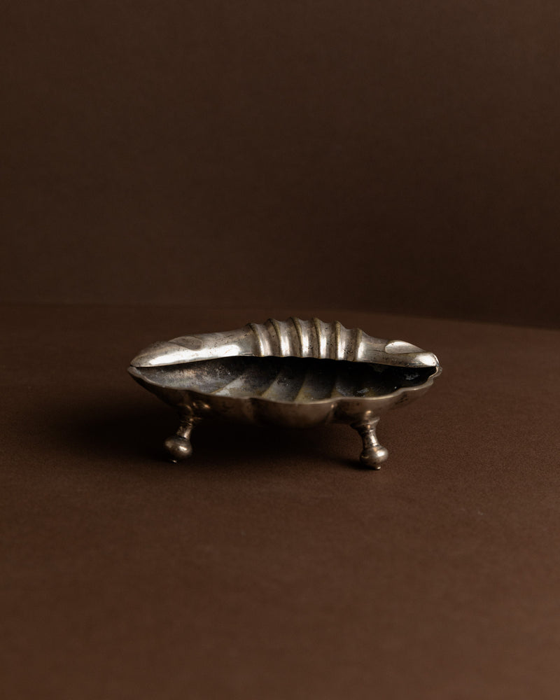A small vintage ashtray from Bottega Jacobs, designed in a shell shape with three dainty legs, set against a dark brown background. Crafted in Italy during the 1960s, this decorative piece features slightly tarnished silver that exudes timeless charm.
