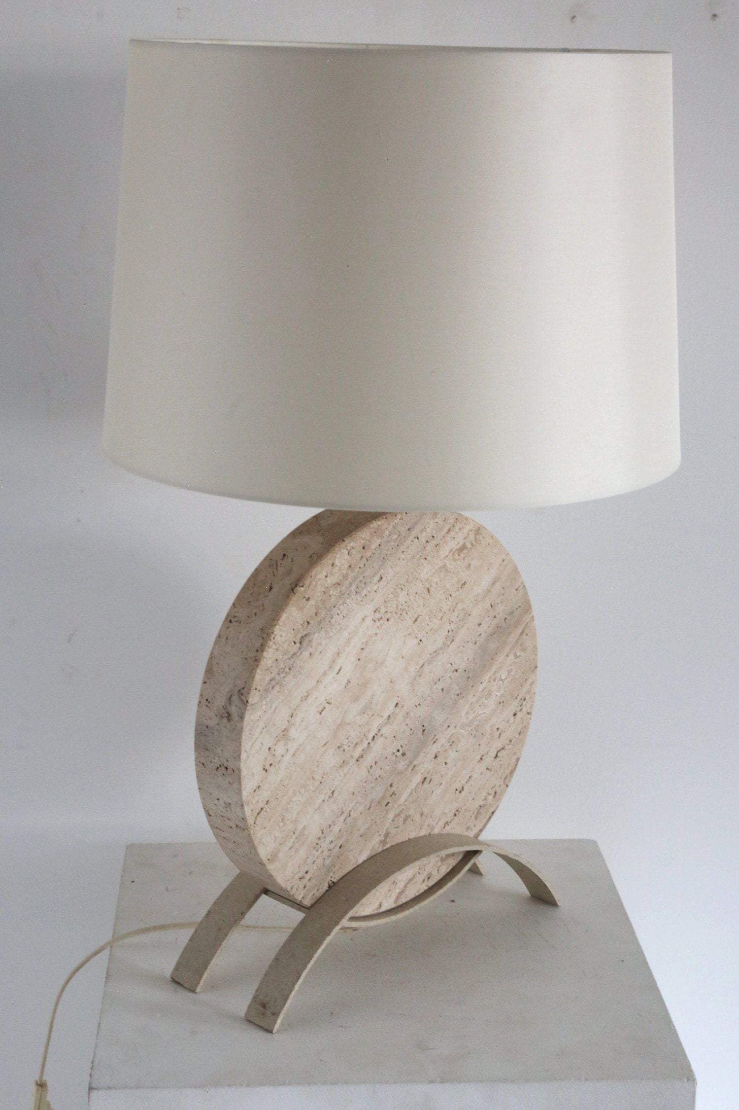 The Italian Travertine Table Lamp by Ancien et Jolie is a beige design with a round marble base, white cylindrical shade, and two curved metal legs on a white surface. A power cord extends from its sculptural base.