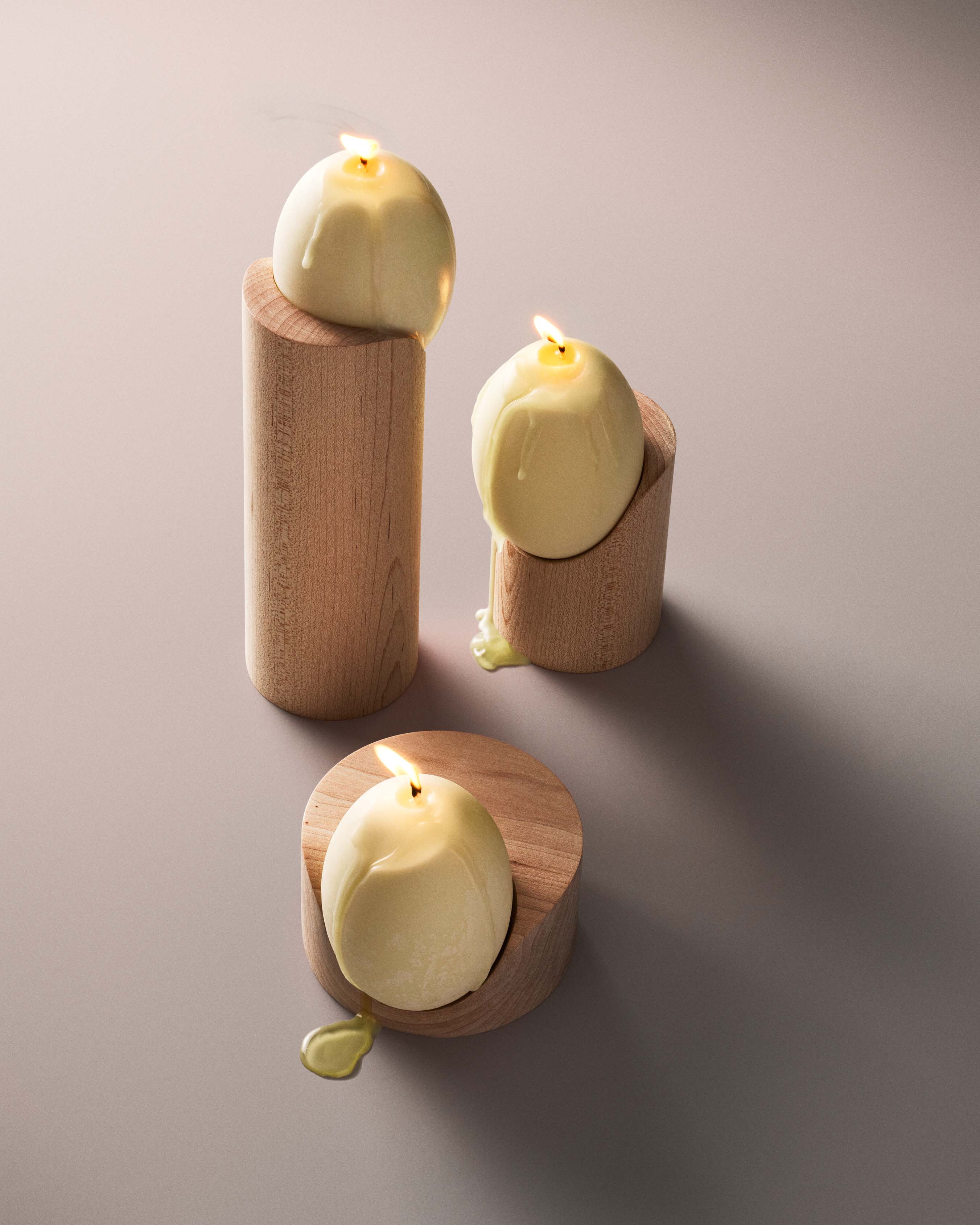 Three lit candles with round yellow bases are placed atop cylindrical wooden stands of varying heights from the Low Perch Candle Holder collection by Tête Studio, reminiscent of Noguchi sculptures. The candles have melted slightly, creating drip marks on the wood. The minimalist arrangement is set against a smooth, neutral background.