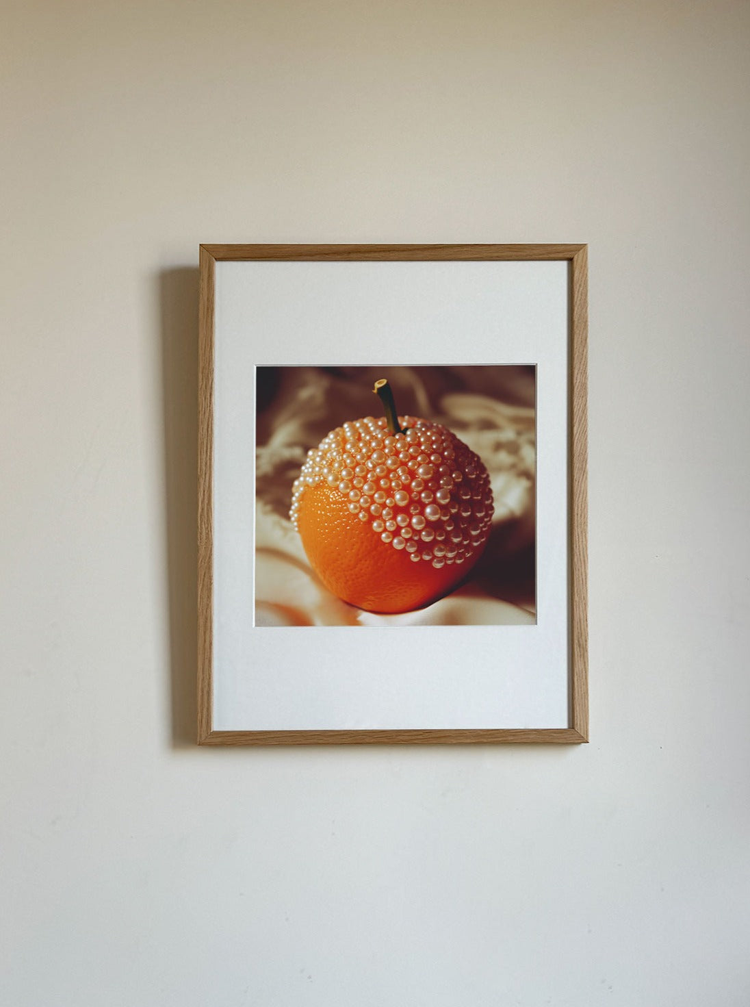 A framed mixed media photography piece shows an orange adorned with numerous small pearls, creating an artistic and striking visual contrast between the natural fruit and the lustrous beads. Orange and Pearls by Adeline Mai boasts a simple wooden frame and is mounted on a plain white wall.