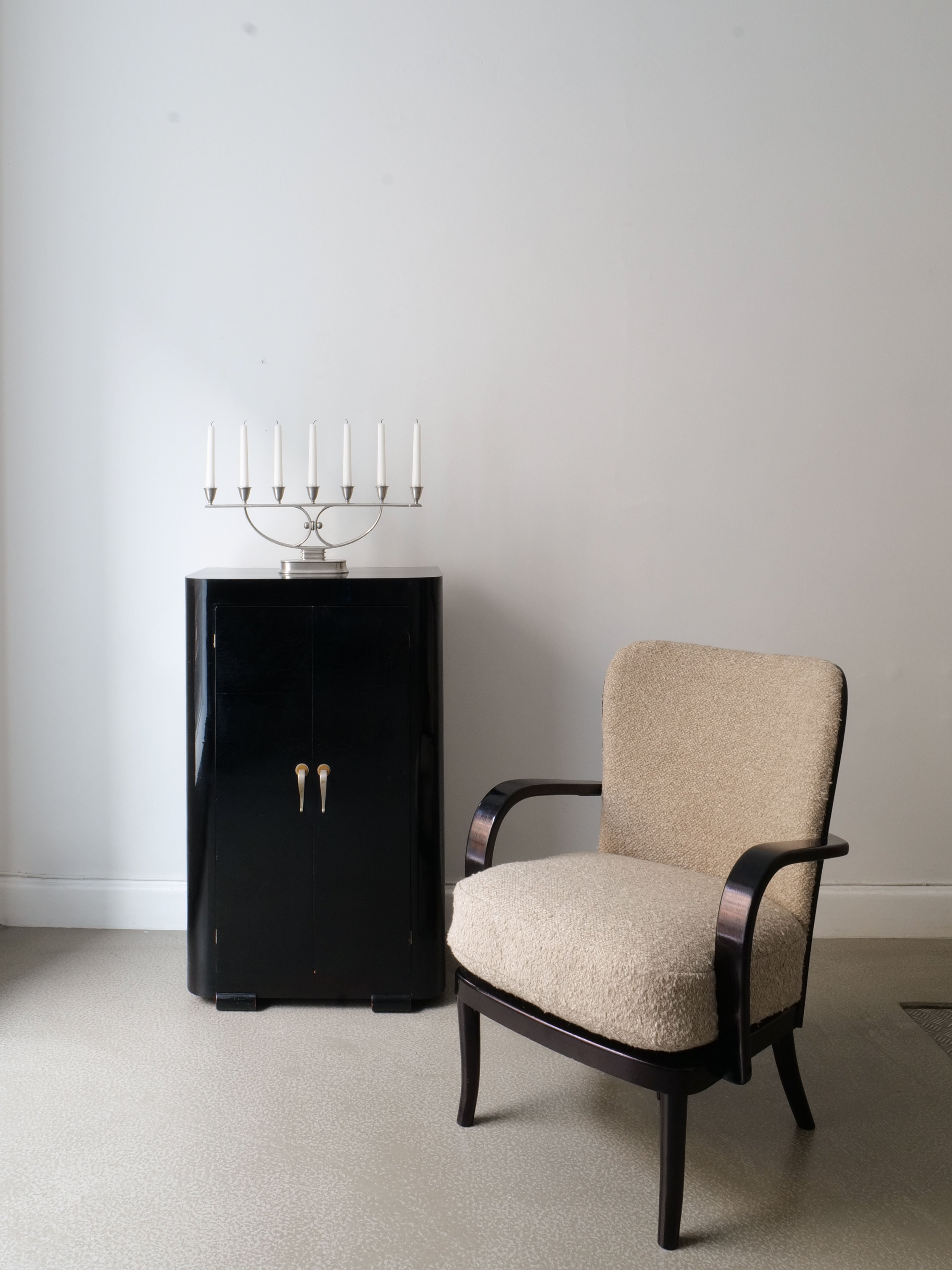 A minimalist room showcasing a beige Pair of Werner West Armchairs from the 1930s by Collection apart, complemented by a tall black lacquered wood cabinet and a silver candelabra with white candles. The backdrop features a plain light-colored wall and a light carpeted floor, creating an elegant setting.