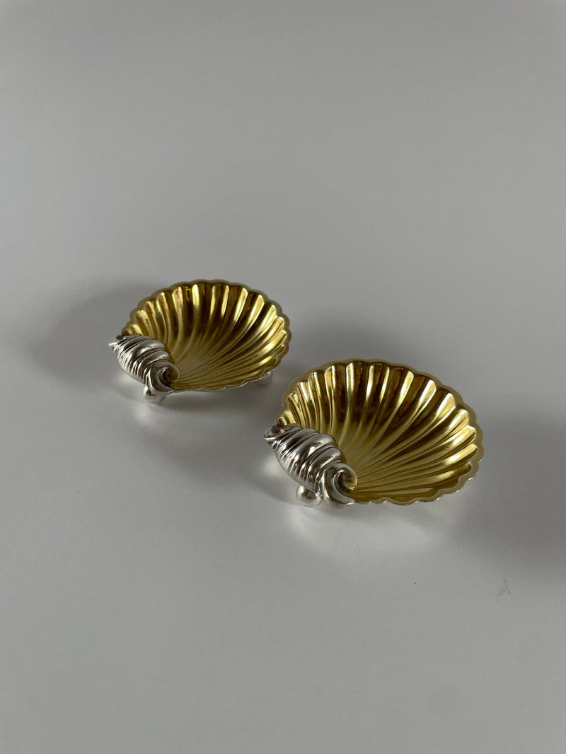 A pair of Maud Vaughan's Vintage Silver Plate Shell Salts features exquisite shell-shaped dishes adorned with intricate ornamental details. Resting on a simple white surface, these decorative elements echo curling sea motifs that evoke the elegance of 1930s decor, highlighting the timeless design of these pieces.