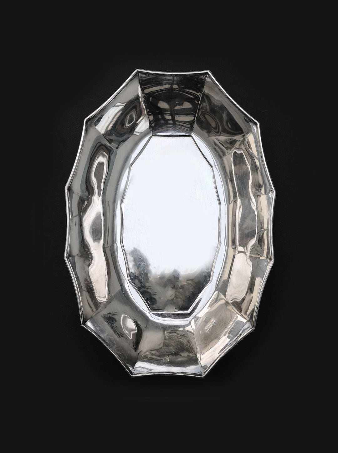 Centered against a black background, the Les Objoies Silver-plated Tray showcases its shiny, reflective finish. This octagonal tray features beveled edges and a smooth, polished surface that exudes sophistication through its intricate reflections.