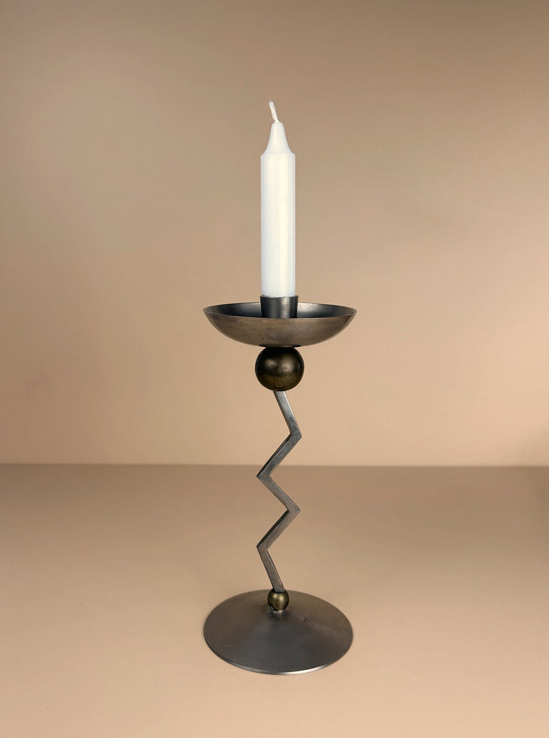 A sleek, modern Postmodern Zig Zag Candle Holder by Dodo Vintage, featuring a silver-colored metal stem and circular base, is centered in the image. It holds a tall, tapered white candle against a soft, warm beige background, evoking Memphis-style design elements.