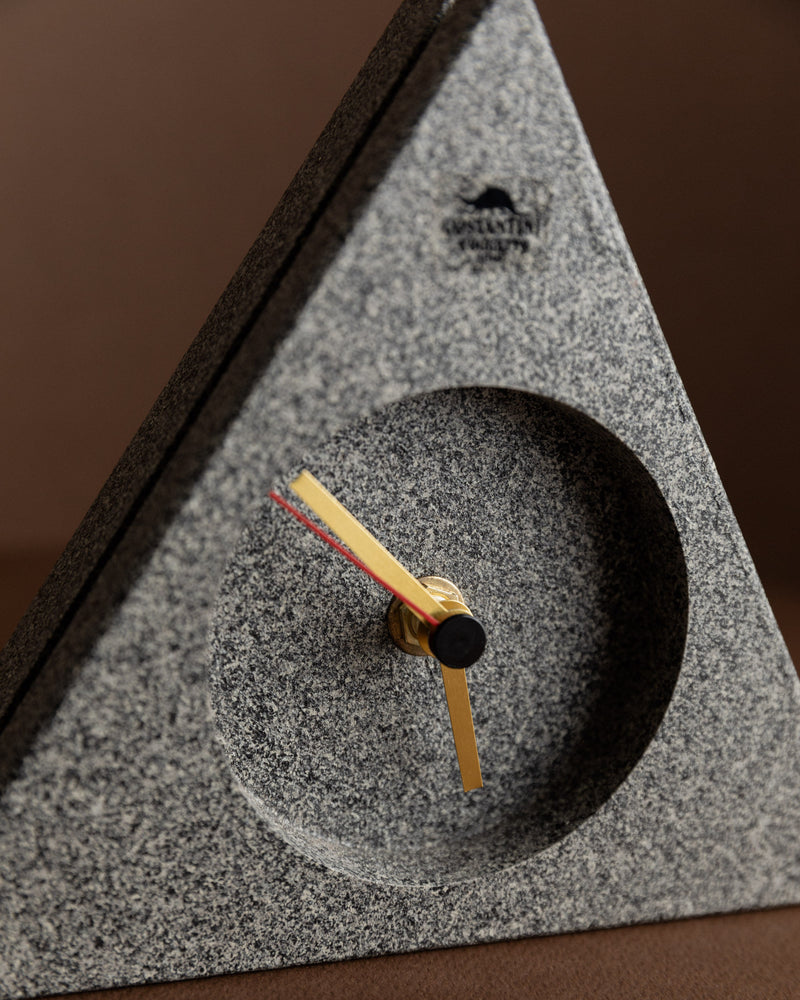 The Vintage Costantini l'Oggetto grandfather clock from Bottega Jacobs showcases a triangular design with a minimalist touch, infused with postmodern Memphis style. It features gold hour and minute hands, a red second hand, and rests on a brown surface. A subtle engraved logo near the top edge completes this unique piece, making it an ideal retro gift idea.