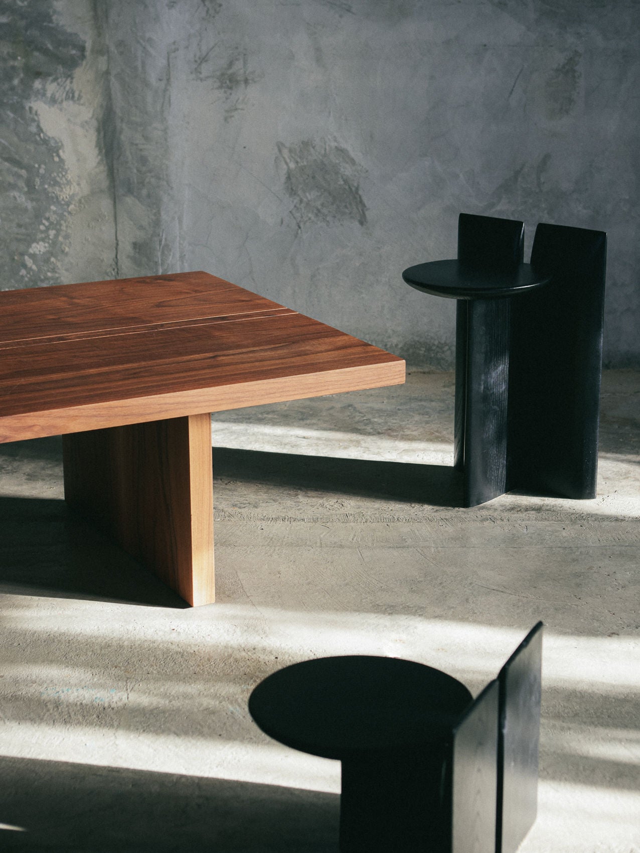 A modern interior showcases a square wooden table with thick legs, paired with two black contemporary chairs. The concrete floor and textured walls are enhanced by the Tabi Stand from Obstacles, creating a minimalist, industrial vibe with intriguing geometric elements.