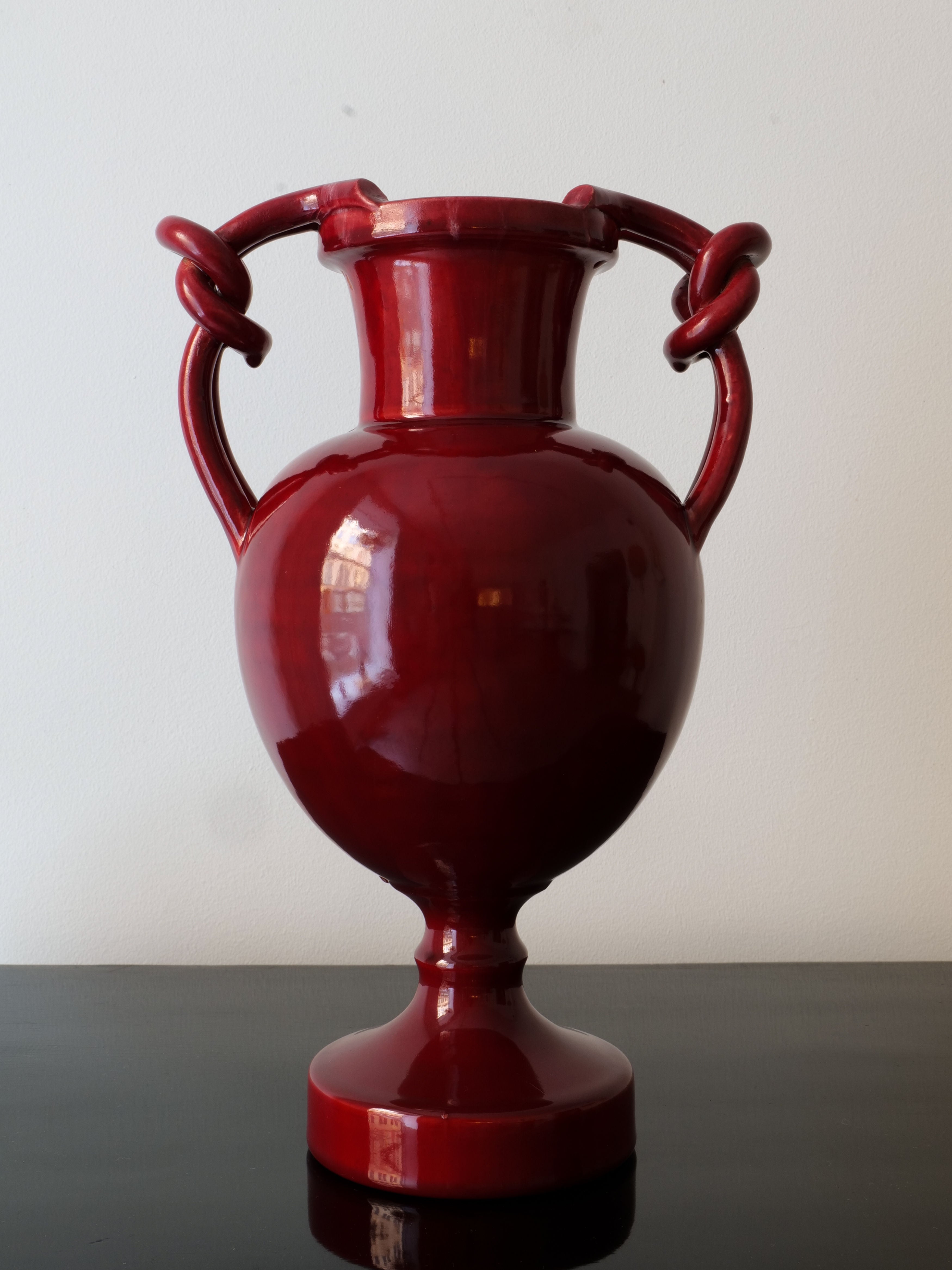 A glossy, dark red Baluster Vase Jérôme Massier by Collection apart is placed on a black surface. Its round body with symmetrical knot-designed handles evokes the French Art Nouveau style, reminiscent of Vallauris ceramics. The piece distinctly stands out against a pale, plain wall.