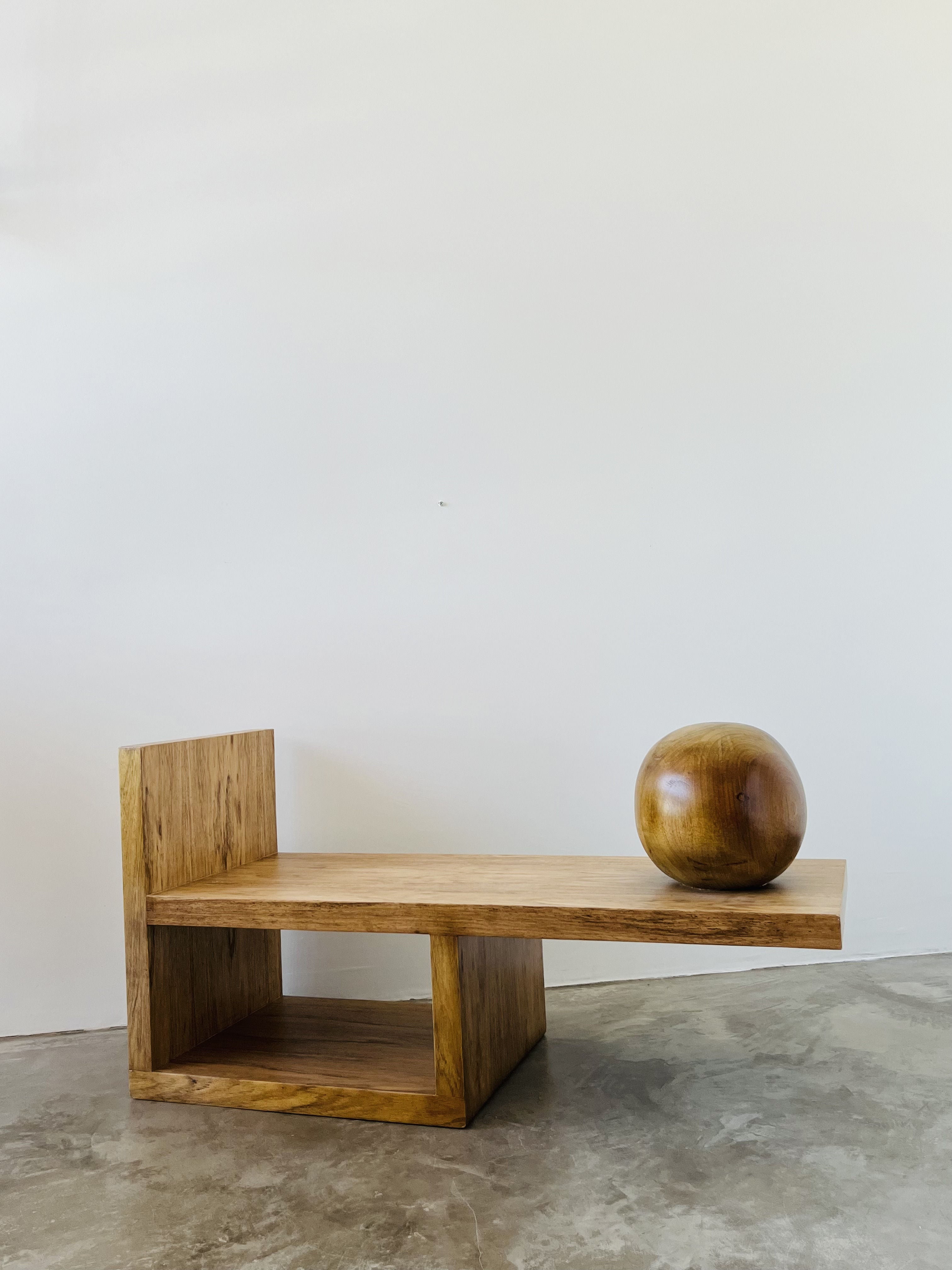 The Lina Side Table by HT. is crafted from Brazilian Freijó wood, showcasing an angular design on a polished concrete floor. A smooth, round wooden sculpture rests atop it, accentuated by a plain white wall that highlights its minimalist aesthetic.