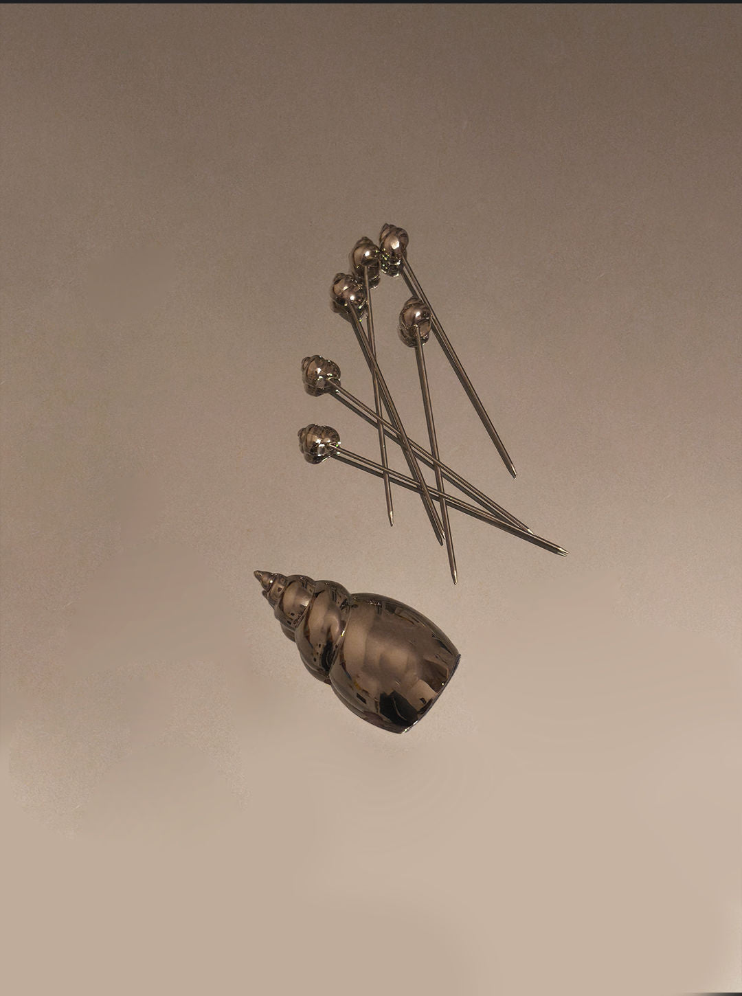 Six metallic Shell Cocktails Picks by Les Objoies, each with a round head, are displayed on a smooth beige surface beside a small decorative shell holder with an ocean-inspired design.