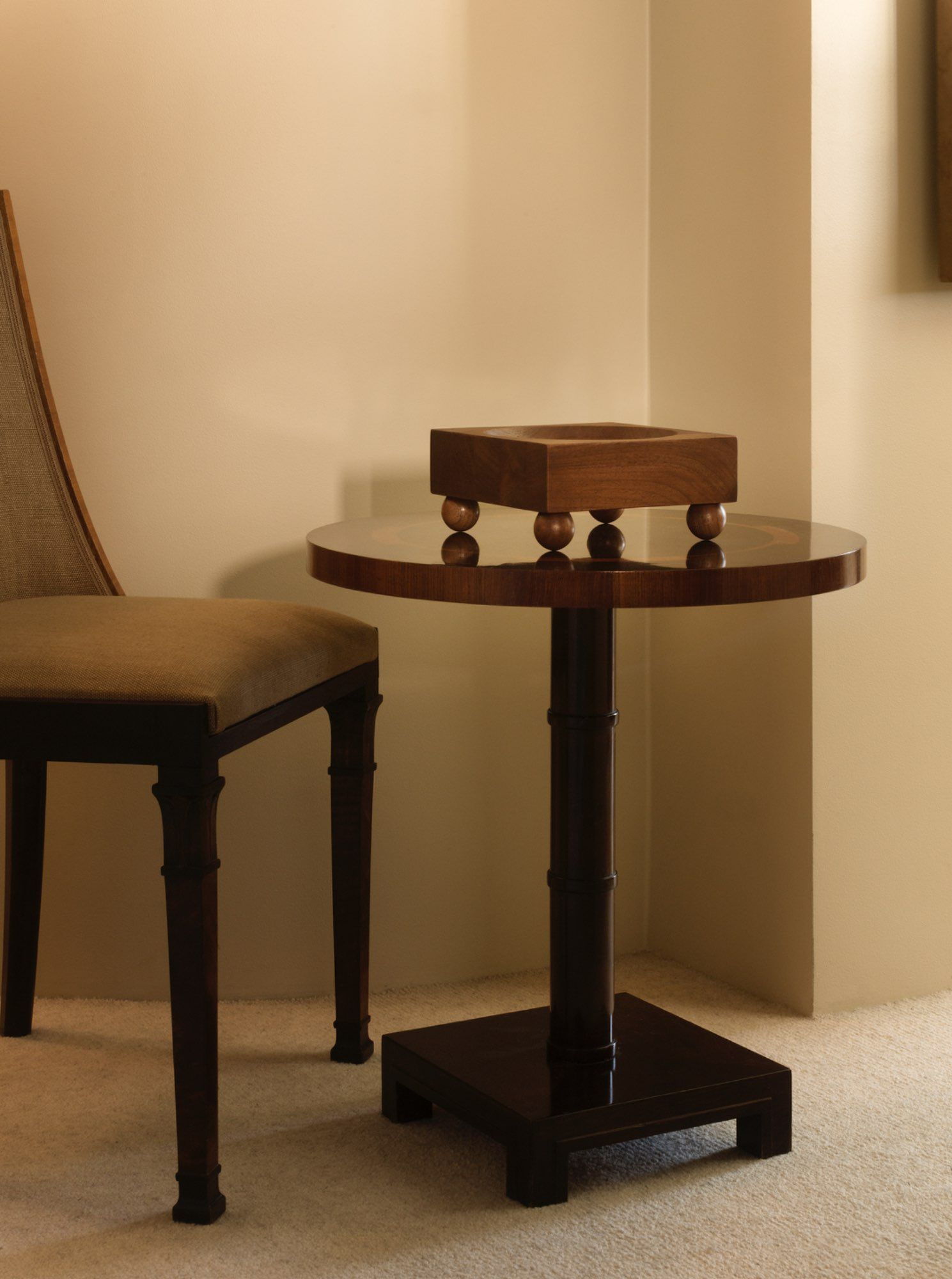 A small, round wooden side table named Tray 01 Walnut from Studio Vraco, featuring a square block decoration on top, sits next to an upholstered chair. The furniture is placed on a light carpeted floor against a beige wall, creating a cozy and minimalist setting.