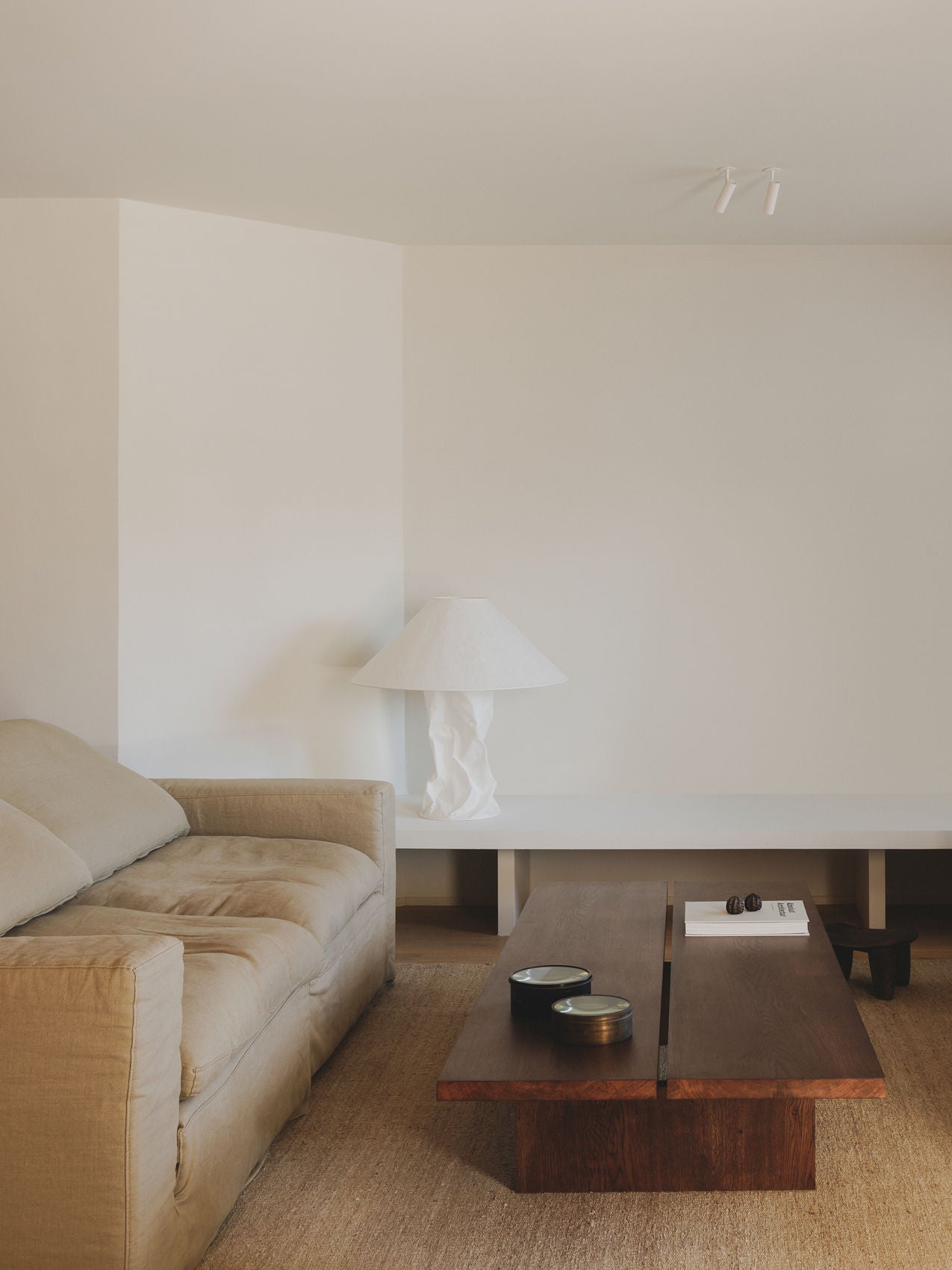 A minimalist living room features a beige sofa, the Tanizaki Coffee Table by ASERIES OF OBSTACLES, SL, and a white sculptural lamp on a bench, emphasizing natural materials and neutral tones for a calm, inviting atmosphere reminiscent of Japanese design.