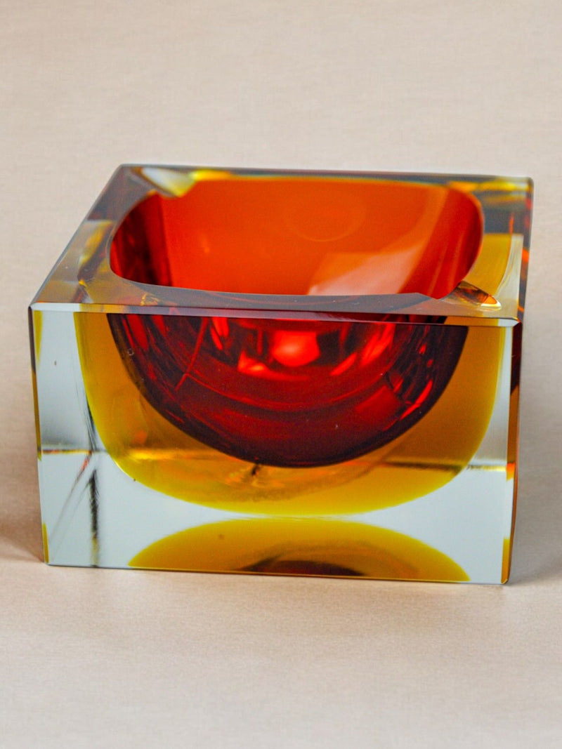 This collectible decorative piece, known as the Sommerso Ashtray by Seguso from the Murano 70s collection by Malamar Studio, is a cube-shaped glass ashtray featuring the Murano sommerso technique with a vibrant orange interior and clear exterior, elegantly displayed on a beige background.