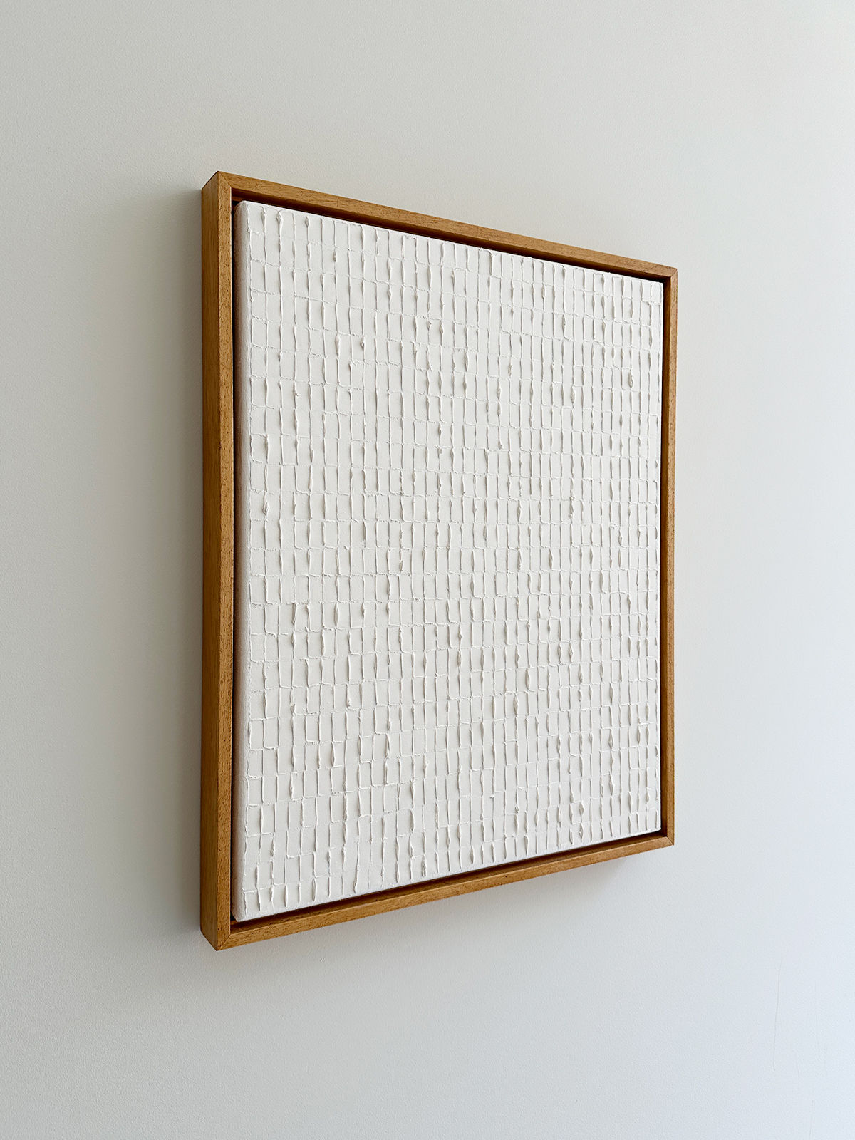 A framed "Warm White - Oil on canvas" by Roi Elmaliah, evocative of the Korean Dansaekhwa movement, consists of a grid of tiny vertical lines and raised rectangular shapes. The wooden frame subtly contrasts against the light gray wall, creating a minimalist and modern effect that encourages mindfulness.