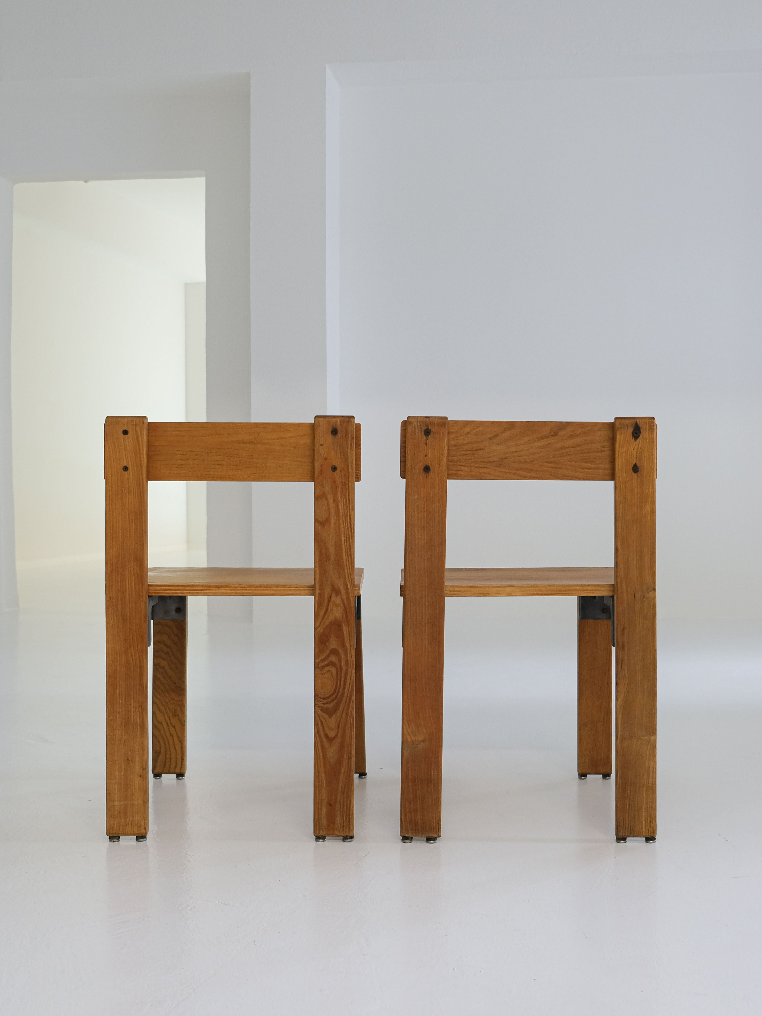 Two "Les Carats" Dining Chairs by George Candilis and Anja Blomstedt, crafted from oak plywood, sit on a white floor against a plain white wall. Facing away from the viewer, the room looks bright and spacious with an open doorway in the background. Brand: antibeige.