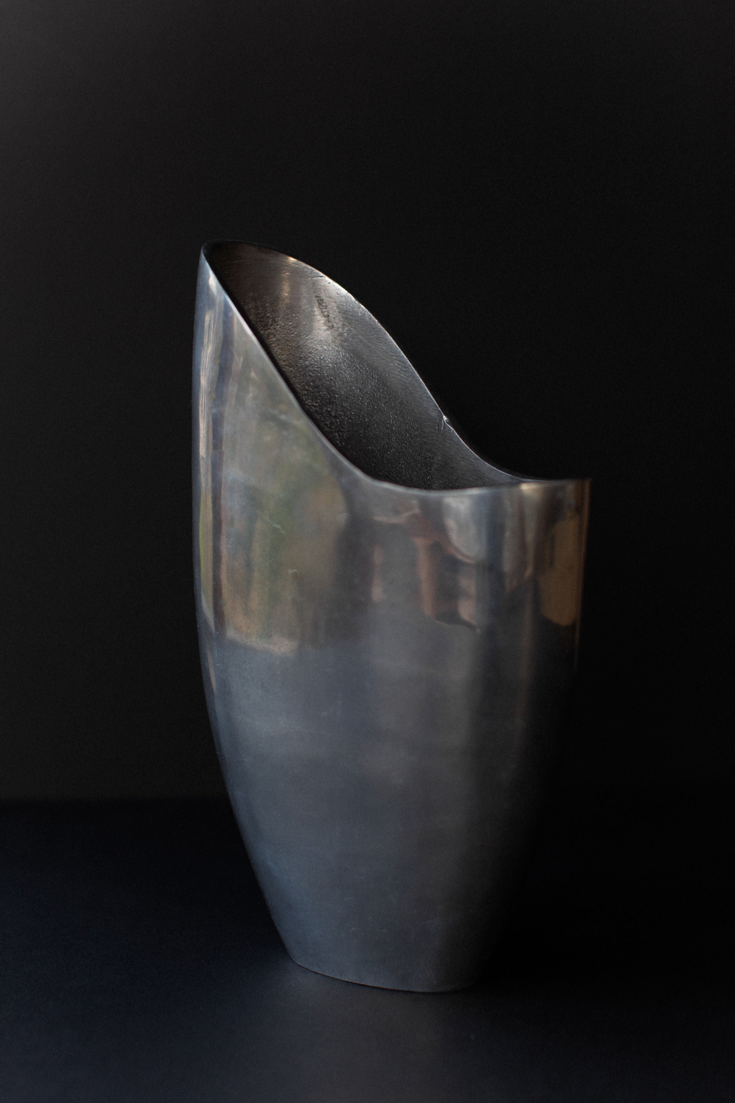 The Out For Lunch Aluminum Cast Vase, with its modern abstract design, boasts a smooth, curved surface and tilted opening. Set against a dark background, it reflects light and casts subtle shadows on its shiny exterior, highlighting its sculptural presence.