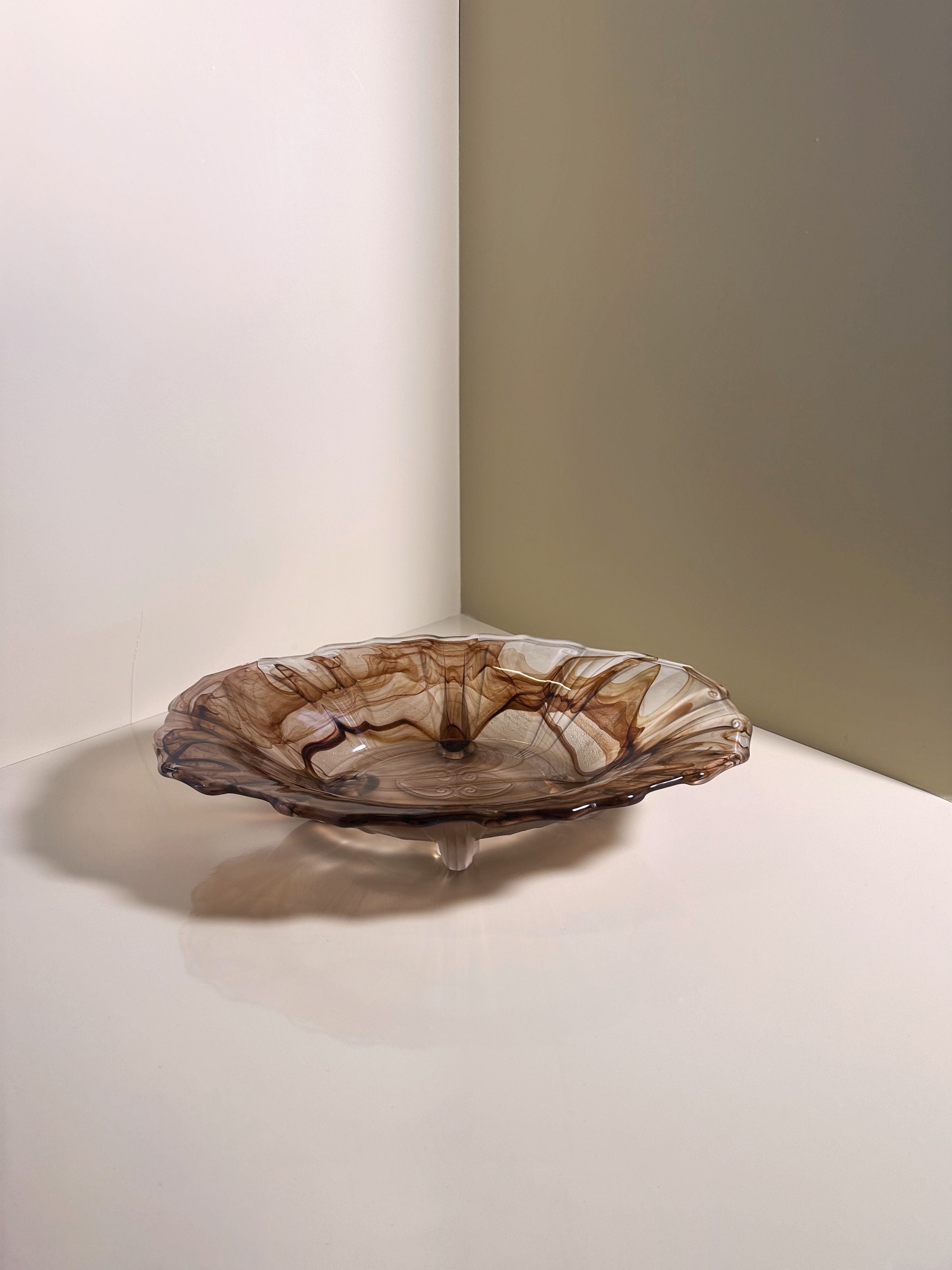 A German Art Deco Cloud Glass Bowl by Dodo Vintage from circa 1930, featuring wavy edges and a brown smoky pattern like Walther Glas, sits on a glossy surface. The light casts gentle shadows against a soft, neutral-colored wall in the background.