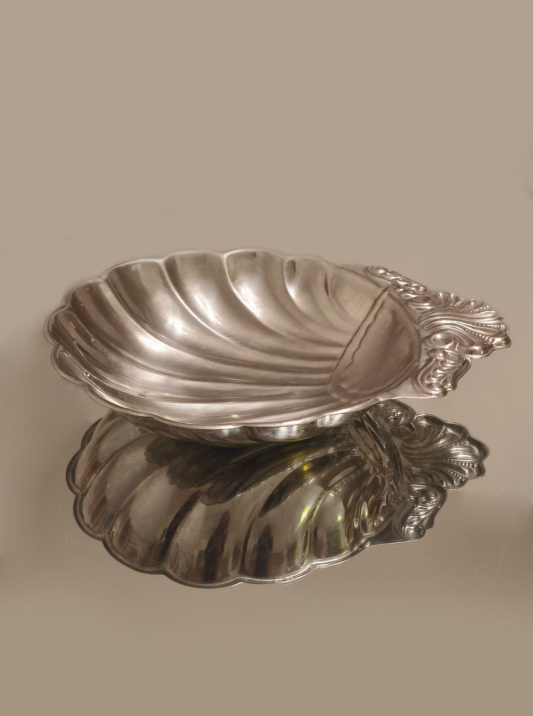 The Large Shell Dish by Les Objoies is a vintage silver piece featuring an ornate handle, elegantly reflecting on a shiny, mirrored surface. Its timeless charm allows it to seamlessly integrate into any setting.