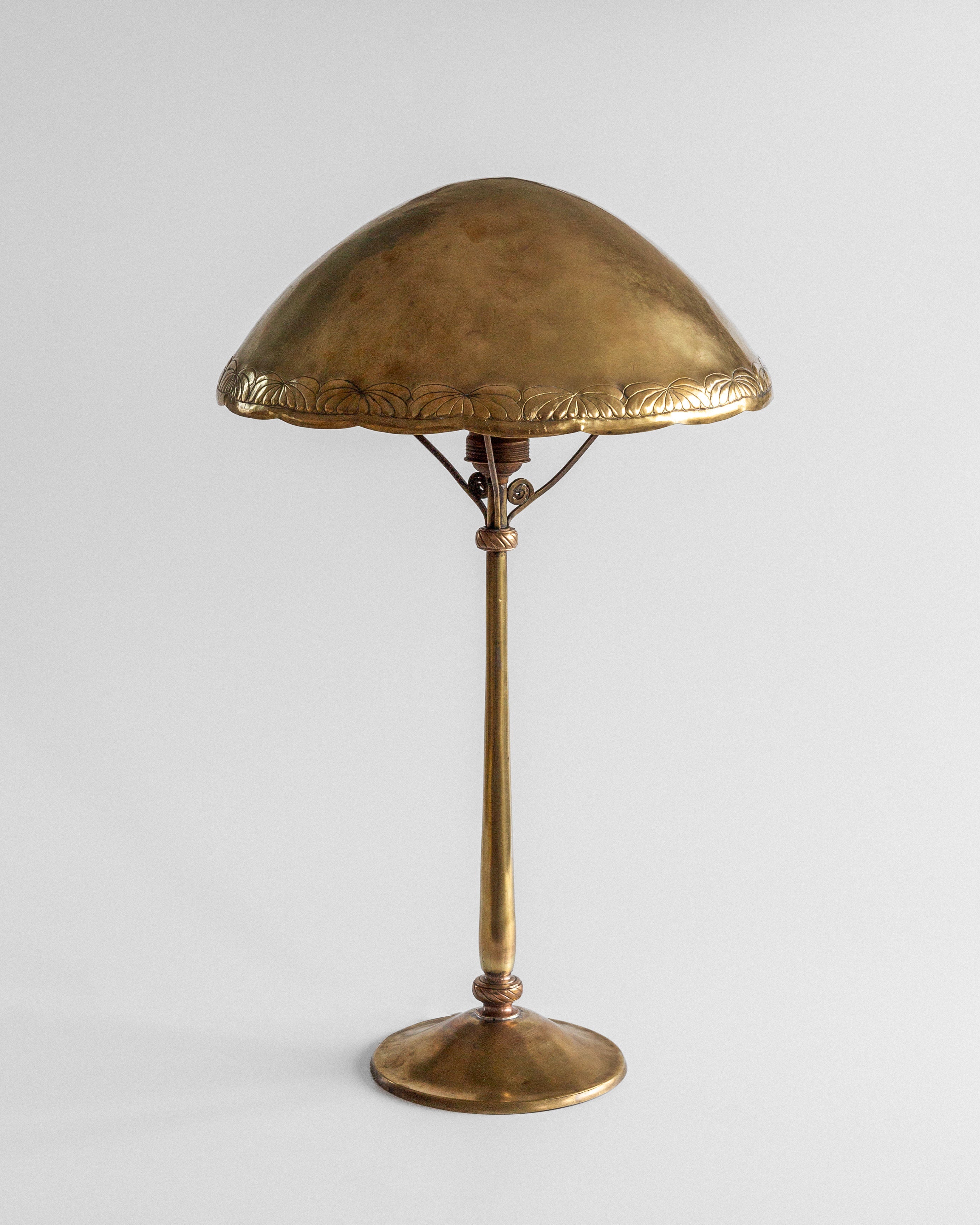 The Spigel Art Nouveau Brass Table Lamp features an ornate, dome-shaped shade with floral engravings and a slender stem on a round base, all set against a plain white background.