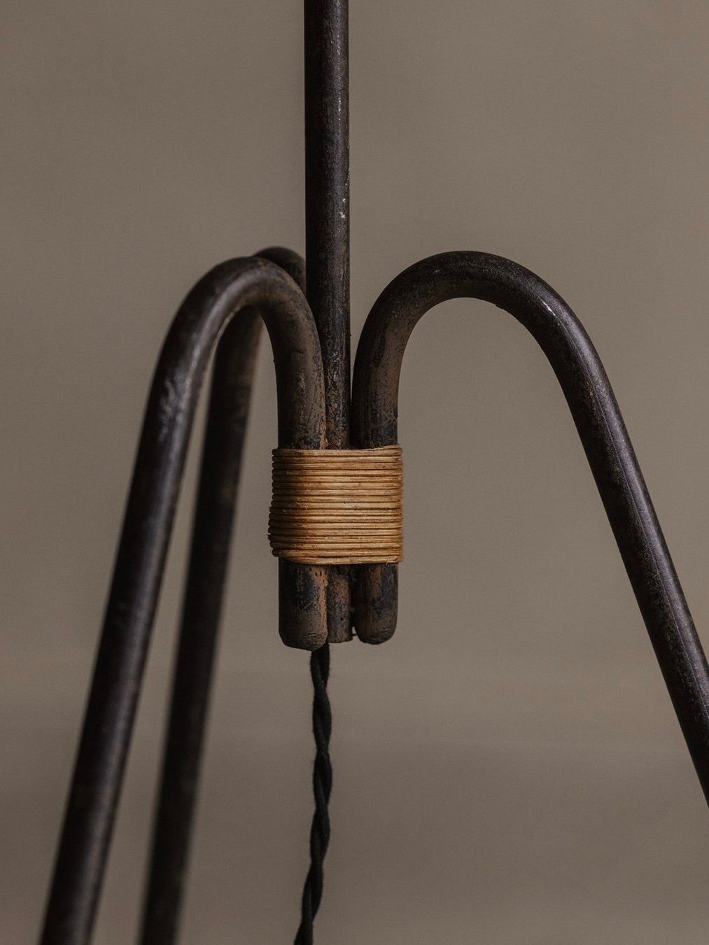 Close-up of the "Manner Of Royere & Adnet" wrought iron tripod lamp by Bicci de' Medici, featuring three legs joined with tan string. A twisted black cable hangs from the center against a neutral background.