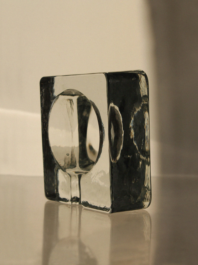 The Brutalist Massive Glass Ashtray Bowl by Vintage Stories sits on a reflective surface, its circular indentation casting soft shadows. Warm light and a neutral background create gentle highlights and 70s-inspired reflections on the glass.