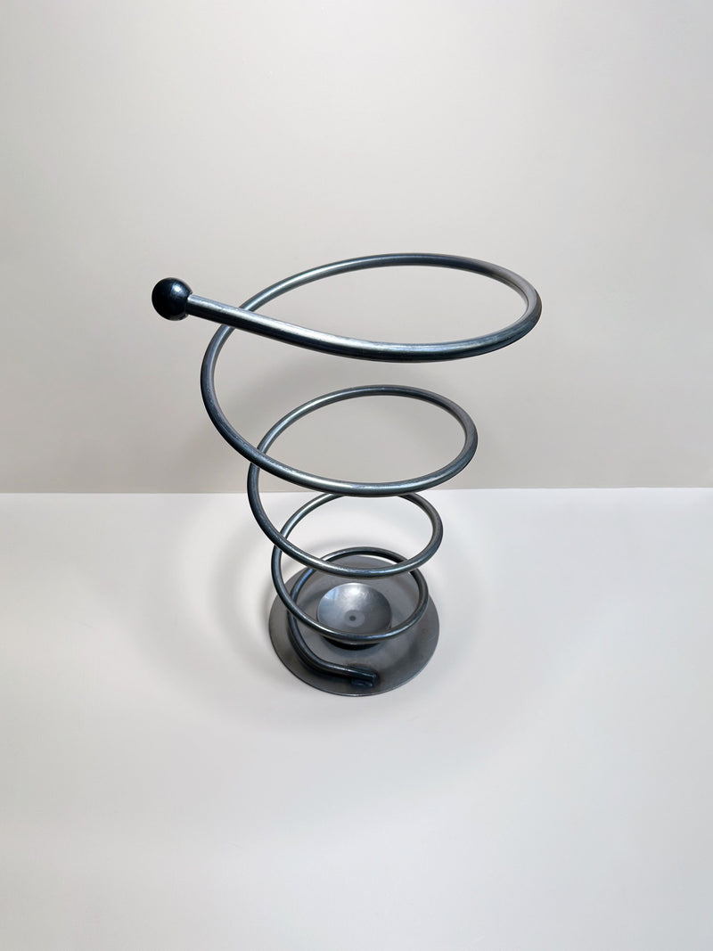 The Dodo Vintage Spiral Metal Umbrella Stand, with its twisted, spiral-shaped structure and circular base reminiscent of a coiled spring, sits elegantly on a flat surface against a plain, light-colored background. This solid metal stand features an elongated loop at the top and exudes a minimalist design that evokes the aesthetic of post-modern decor.