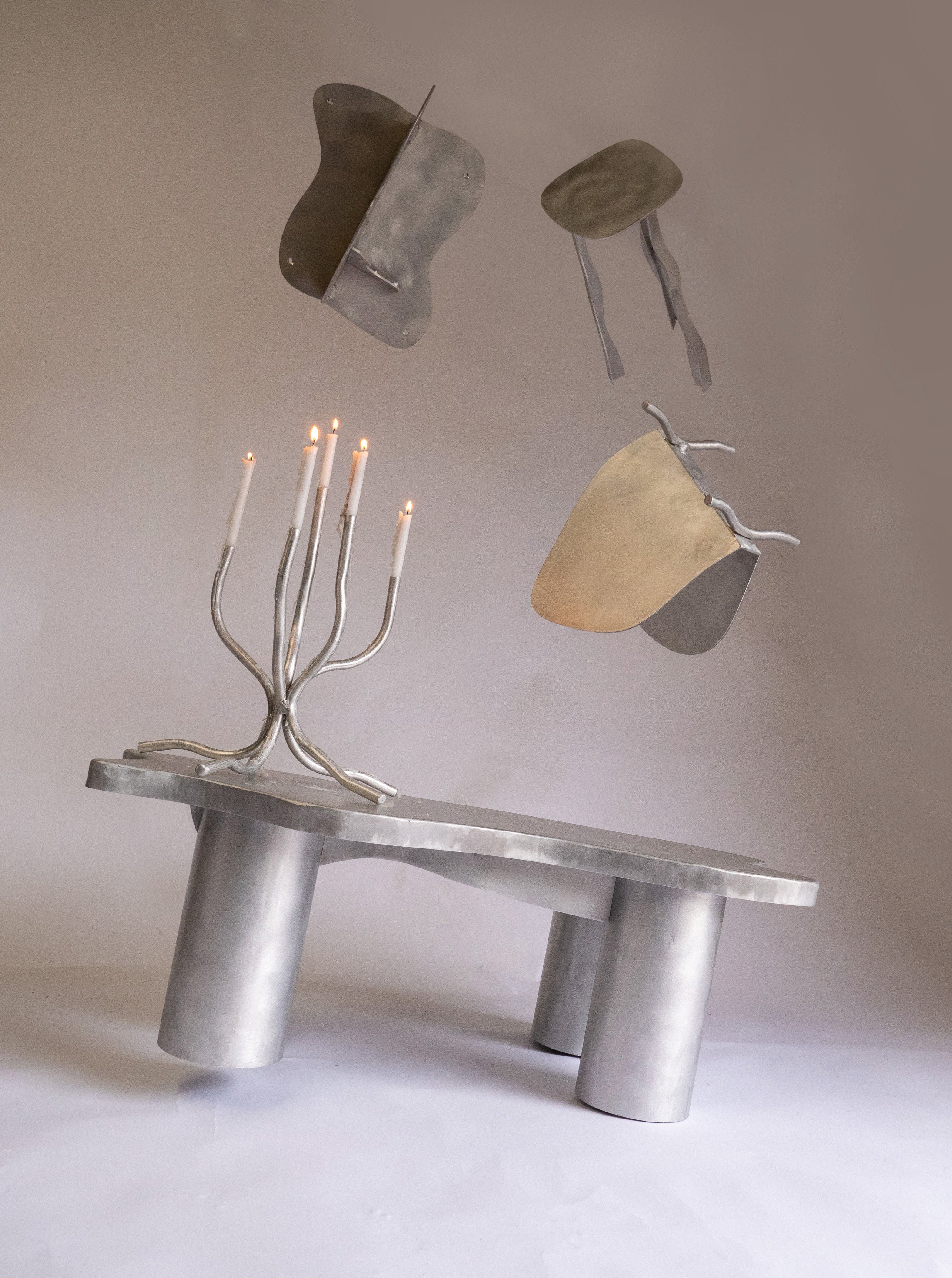A surreal image of a silver bench with a Six Dots Design small candelabra holding lit candles, accompanied by floating, abstract metallic shapes against a soft gray backdrop.