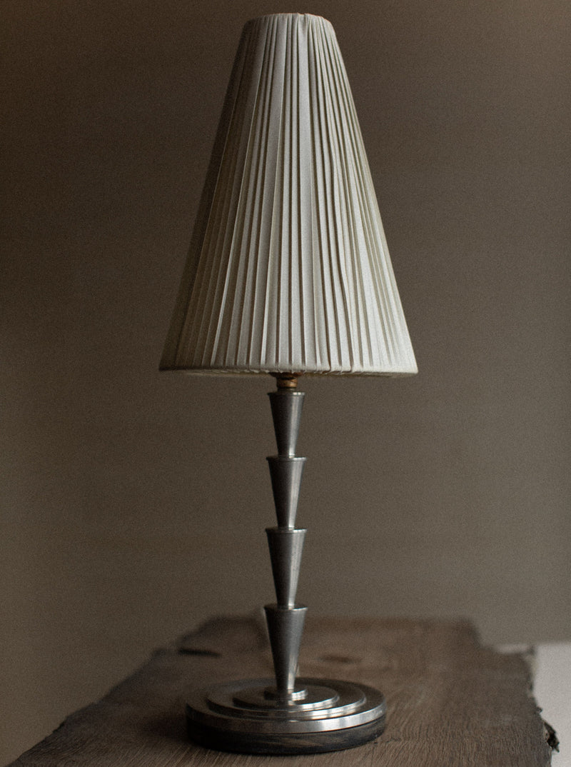 A metallic 1920's Swedish Grace Lamp from Out For Lunch with a pleated, cream-colored shade stands on a wooden surface. The Elis Bergh design features a tiered, conical base and emits a warm, muted light. The background is slightly blurred and neutral in color, enhancing the vintage product’s elegance.