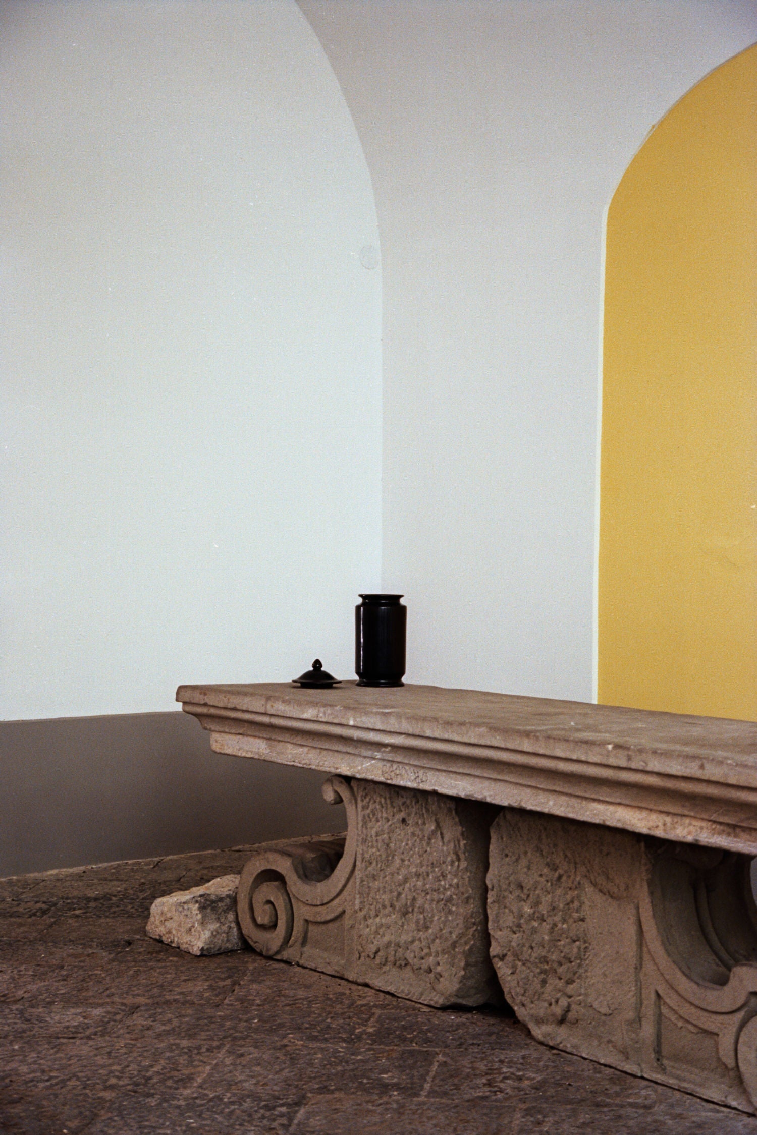 A stone bench with ornate supports stands by a wall of white, grey, and yellow. Atop the bench sits COSEINCORSO’s "Albarello" Jar, a ceramic apothecary piece in matte black, accompanied by a small object for a minimalistic look.