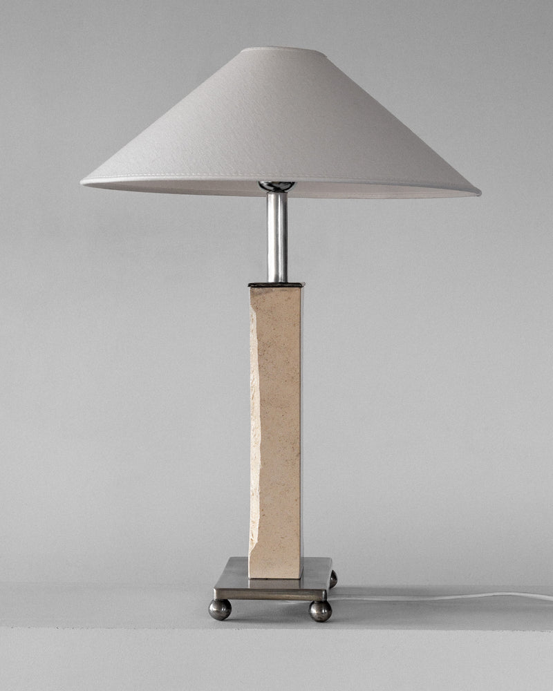 The Spigel Modernist Travertine and Metal Table Lamp features a conical white shade, a stone-textured rectangular base, metallic accents, and is powered by a visible white cord, set against a plain gray background.