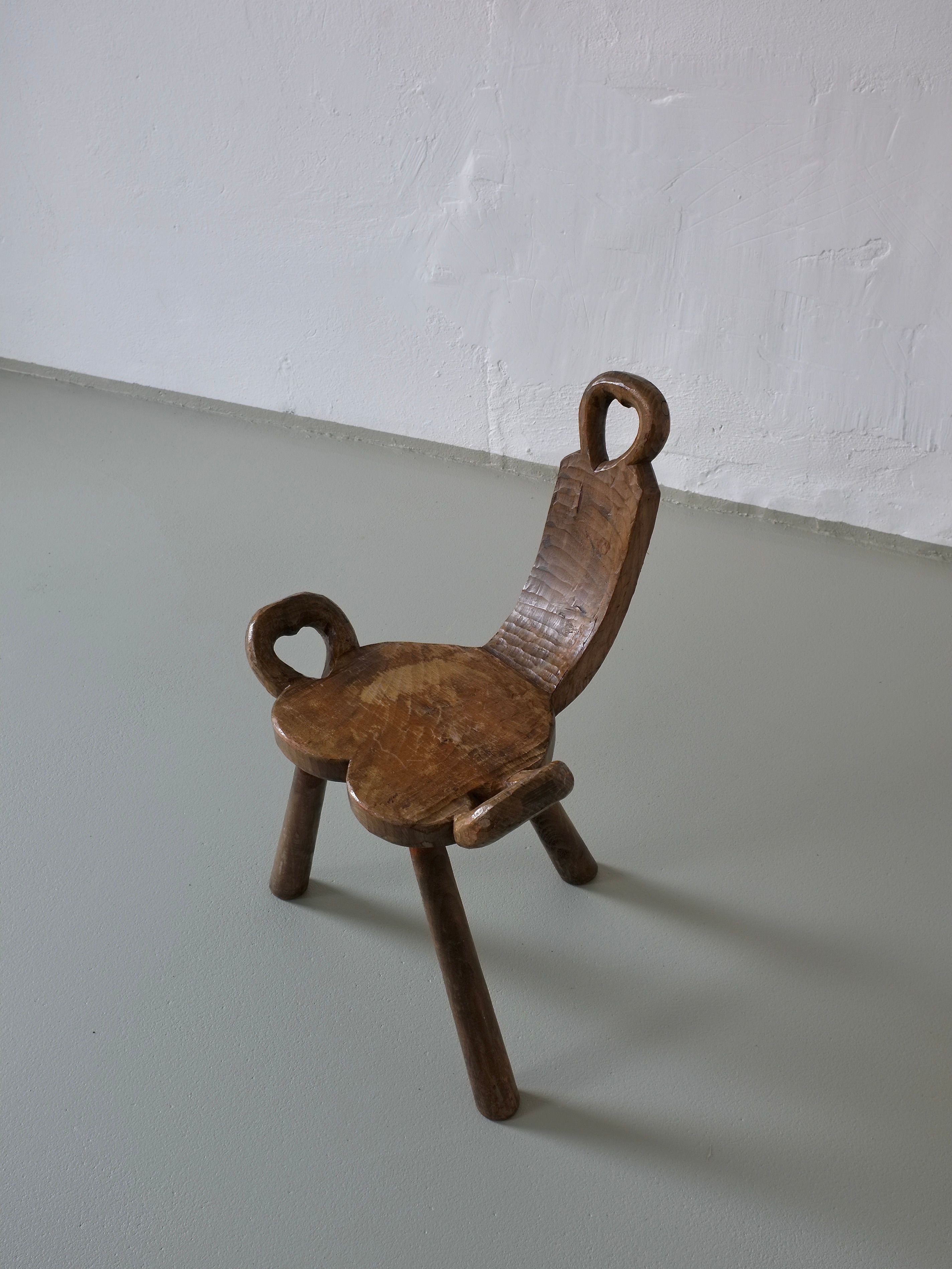 The Vintage Brutalist Carved Tripod Stool by Veter Vintage features a carved backrest and seat with intricate loops on its legs. It's displayed on a smooth, light-colored floor against a plain wall.