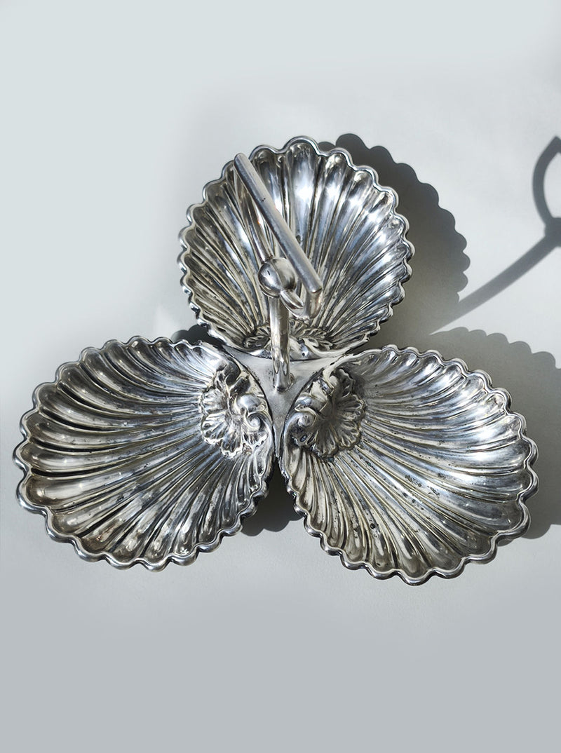 The Les Objoies Large Silver-plated Triple Tray features three shell-shaped compartments arranged in a triangle with a central handle, casting elegant shadows on light surfaces—a timeless serving piece.