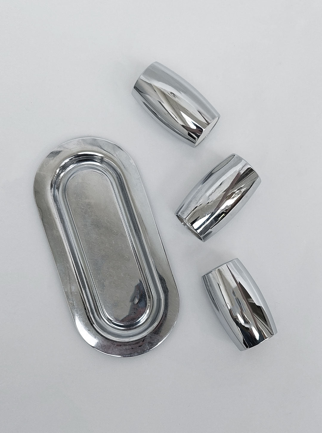 A Salt & Pepper Set from Les Objoies with one large oval piece and three smaller curved pieces, reminiscent of sleek stainless steel. The pieces are placed on a white background, reflecting light off their shiny surfaces.