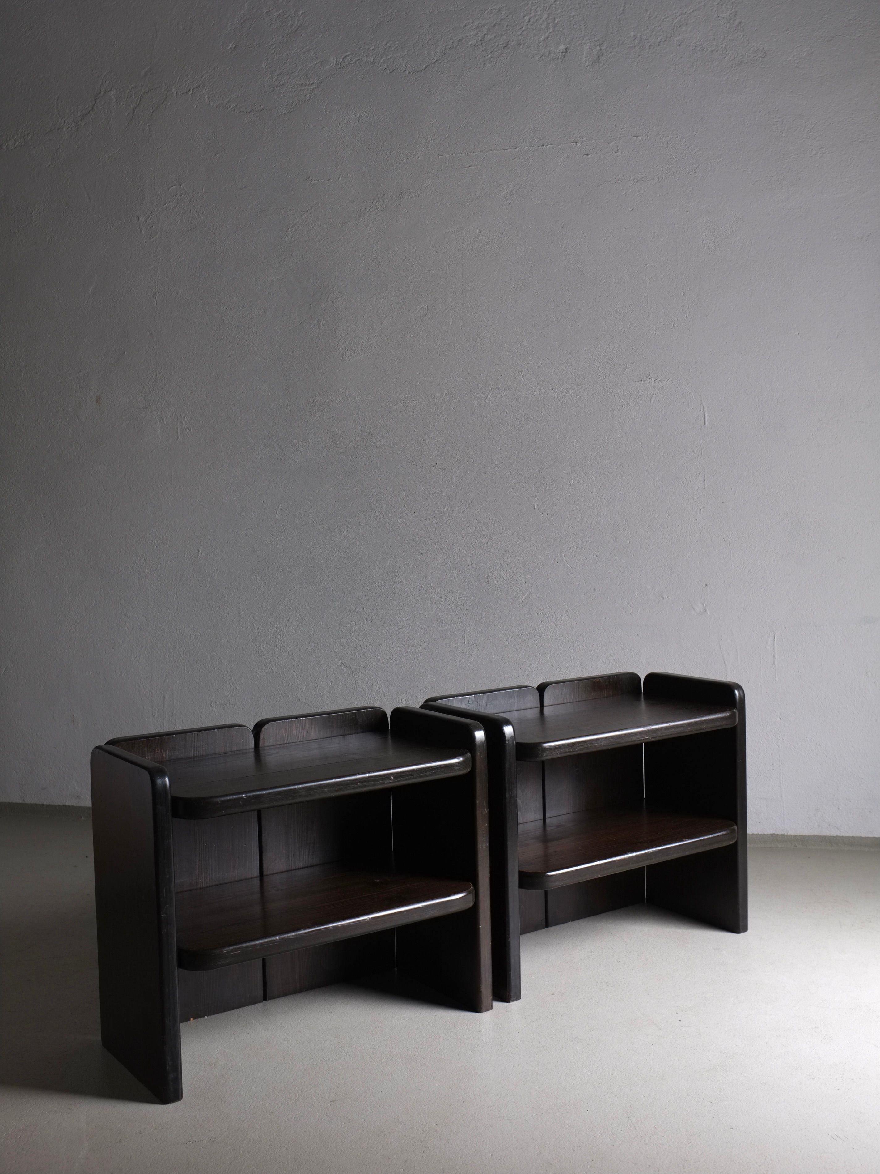 Two sleek, dark wooden 2 Brutalist Dark Wood Night Stands | 1970s by Veter Vintage stand side by side against a plain, light-colored wall. Each nightstand features two shelves with curved edges, showcasing a minimalist, modern design reminiscent of vintage condition. The room's lighting casts soft shadows, highlighting the nightstands' smooth finish.