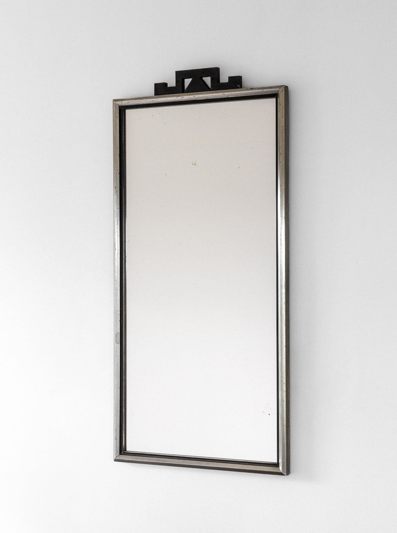 A 1930s Swedish Art Deco mirror by Spigel features a thin, metallic frame and an ornate, black decorative piece on top. The mirror is mounted on a plain white wall and reflects a neutral, empty space.