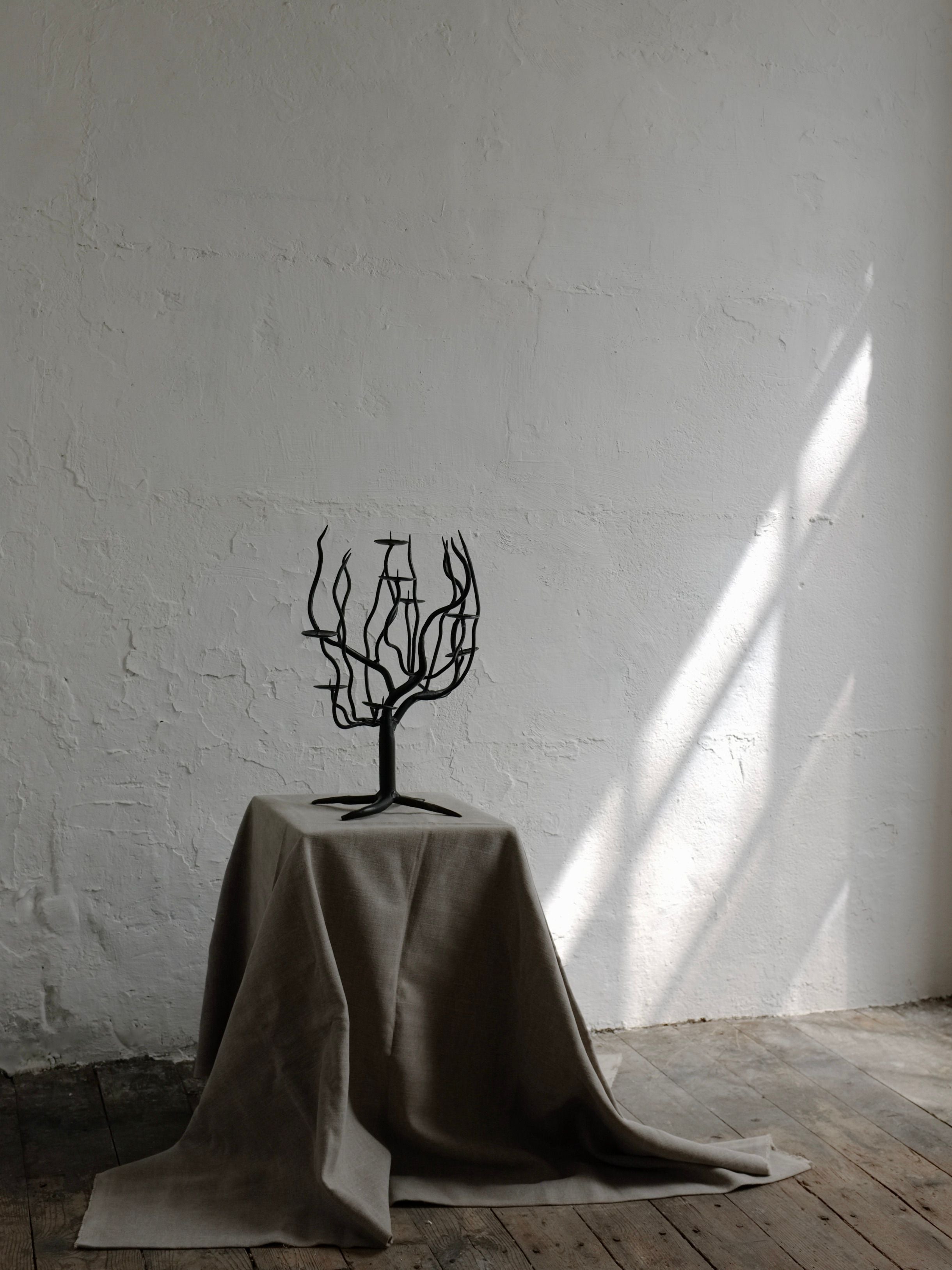 The Hand-forged Metal Sculptural Candle Holder by Veter Vintage, a small black metal piece resembling a tree with bare branches, is placed on a draped beige cloth covering a stand. The stand is set against a textured white wall, and soft sunlight streams through, casting shadows and creating a minimalistic, serene atmosphere evocative of 1960s design.