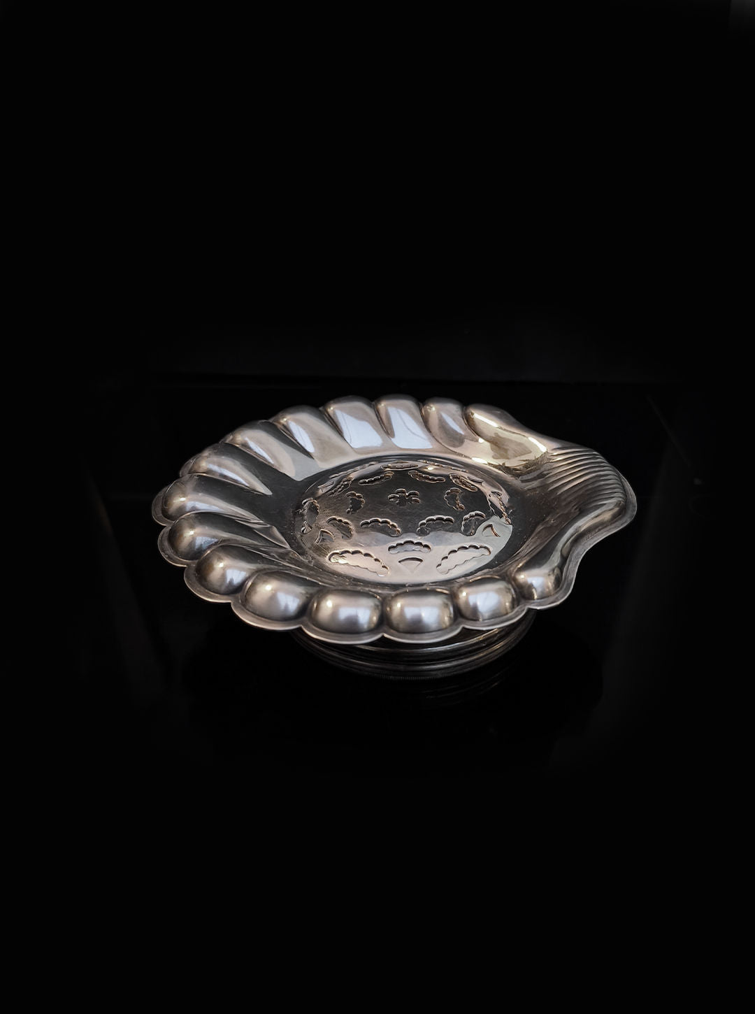 The Shell Butter Dish by Les Objoies features an ornate silver design with a scalloped edge and intricate cutout patterns in the center, reminiscent of culinary art, set against a black background.