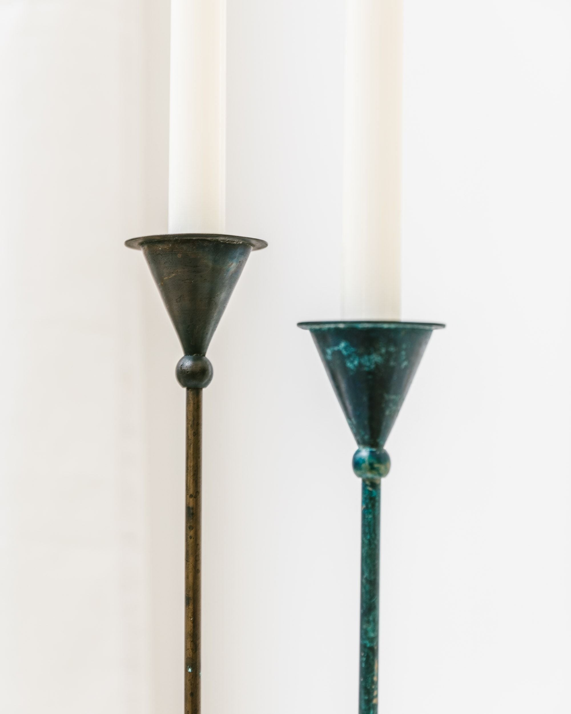 A pair of candleholders from Bottega Jacobs, titled "Set of 2 Candleholders – Postmodern 1980s," feature tall, slender holders with a dark patina finish. Each supports a white candle and stands against a light background, showcasing a conical form reminiscent of vintage Postmodern designs from the 1980s in their simple and elegant design.