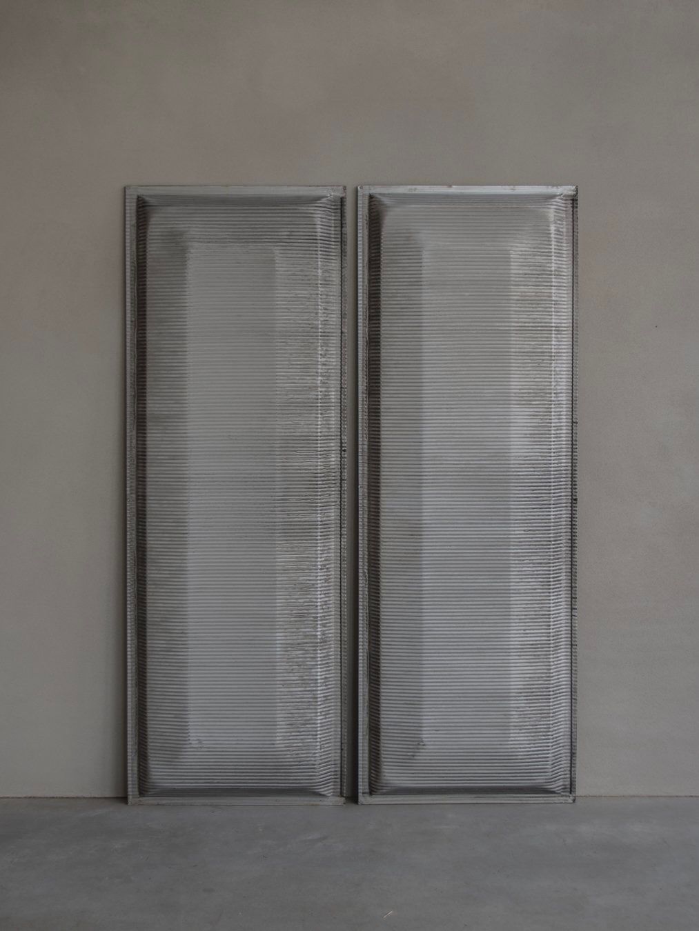 Two large clear rectangular Bicci de' Medici Jean Prouvé - Façade Panel 70 glass panels with metal frames stand side by side against a smooth gray wall. Their slightly textured surfaces subtly reflect light, echoing the modernist aesthetic of Jean Prouvé.