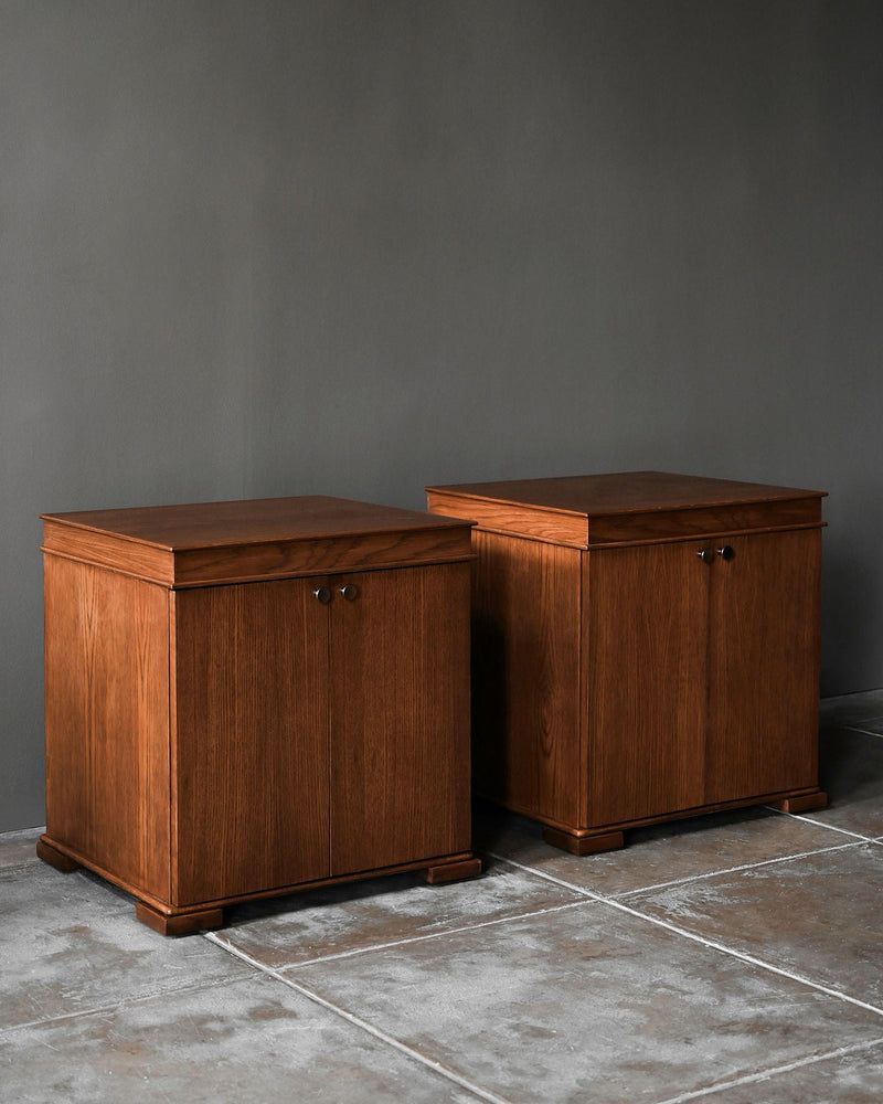 Two Lancaster Nightstands by Studio OSKLO, crafted in dark brown stained oak, sit side by side against a gray wall on tiled flooring. Each handmade piece features two front doors with round handles, showcasing quality design.