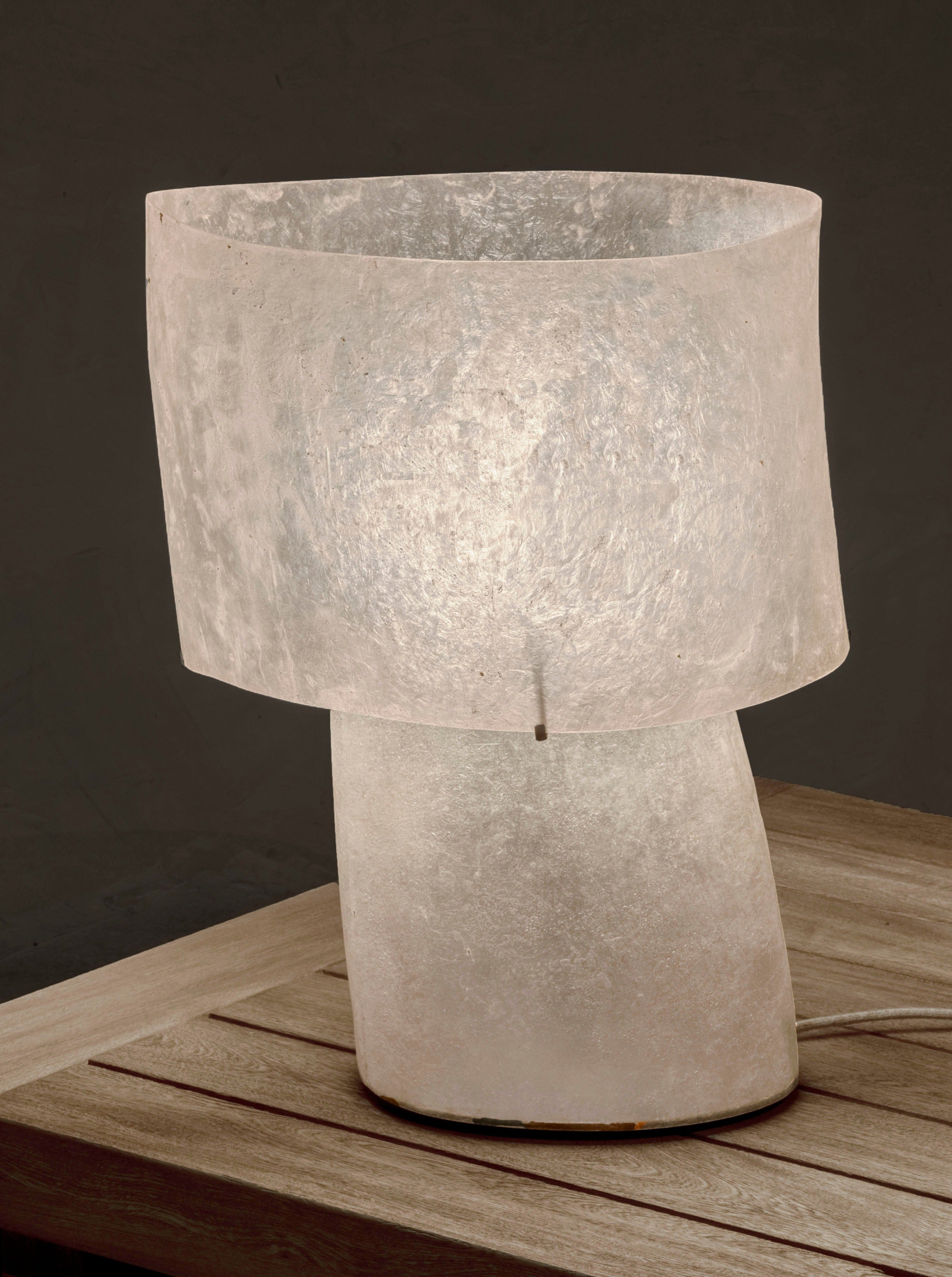 A Kilzi Mush Table Lamp - Tall, with its modern frosted glass design, sits on a wooden table against a dark background. It emulates the mush lamp style, showcasing a cylindrical base and a wide, slightly translucent fibreglass shade that casts an elegant, soft glow.